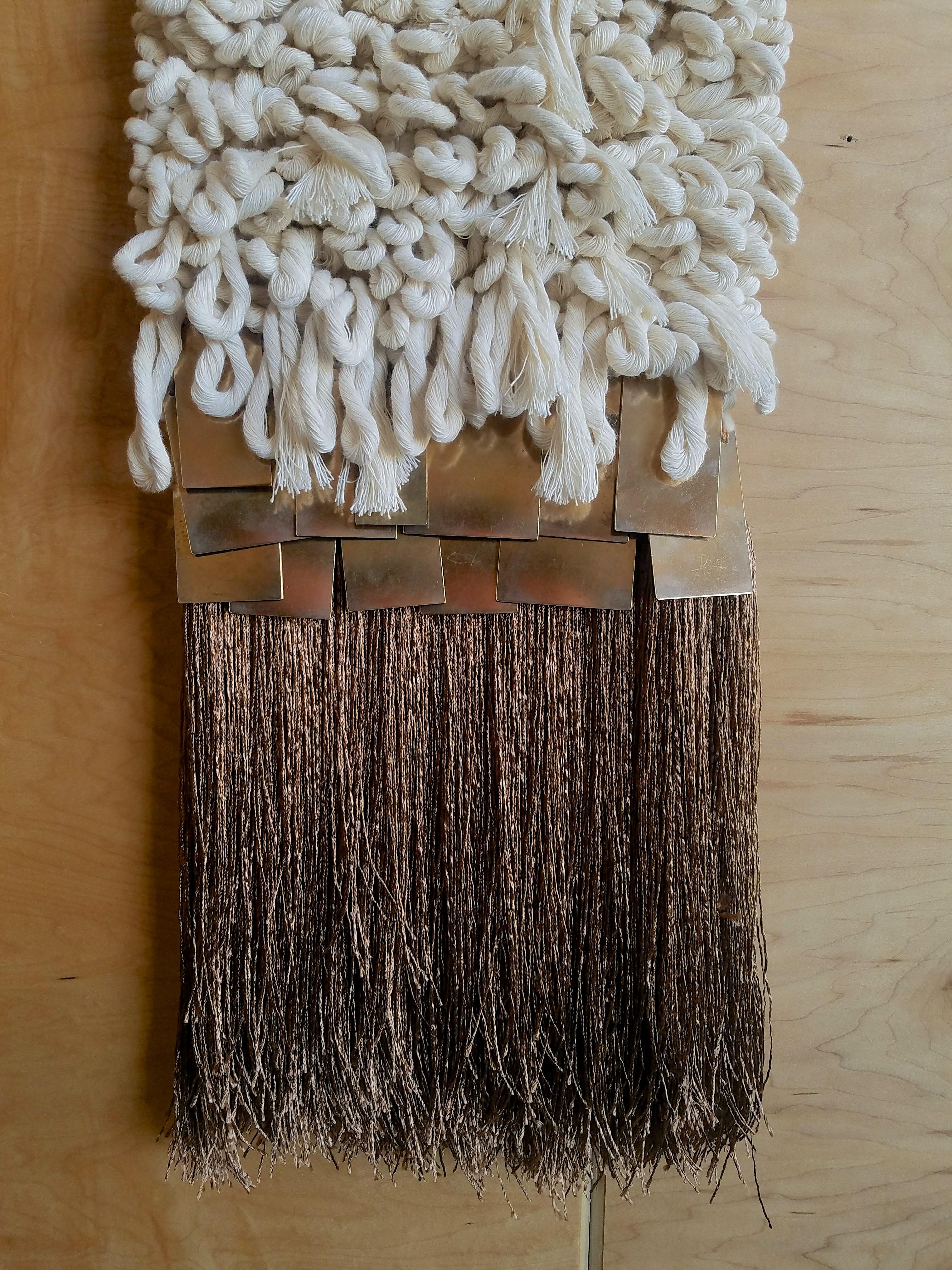 Handwoven, tapestry style, wall hanging by Janelle Pietrzak of All Roads. Rustic tan hemp fiber, natural cream cotton rope and repurposed brass tiles cascade over shiny tassels. Fibers used are hemp, cotton and shiny viscose. Weaving hangs fully