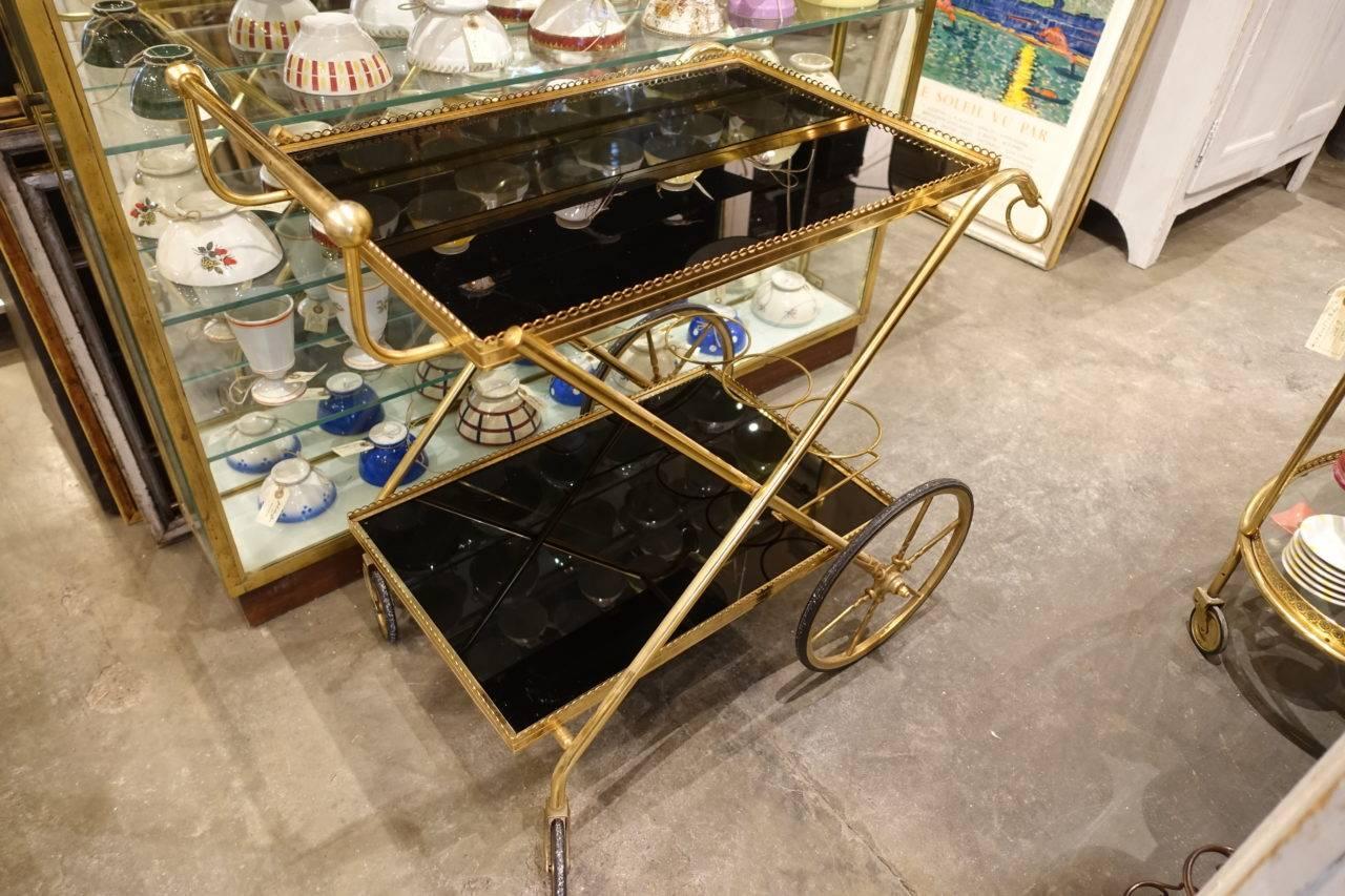 Gorgeous retro French serving trolley with a framework made of brass, two wonderful black glass tops, and two large wheels at the front. Note the beautiful pattern work along the edges. Practical and stylish item.