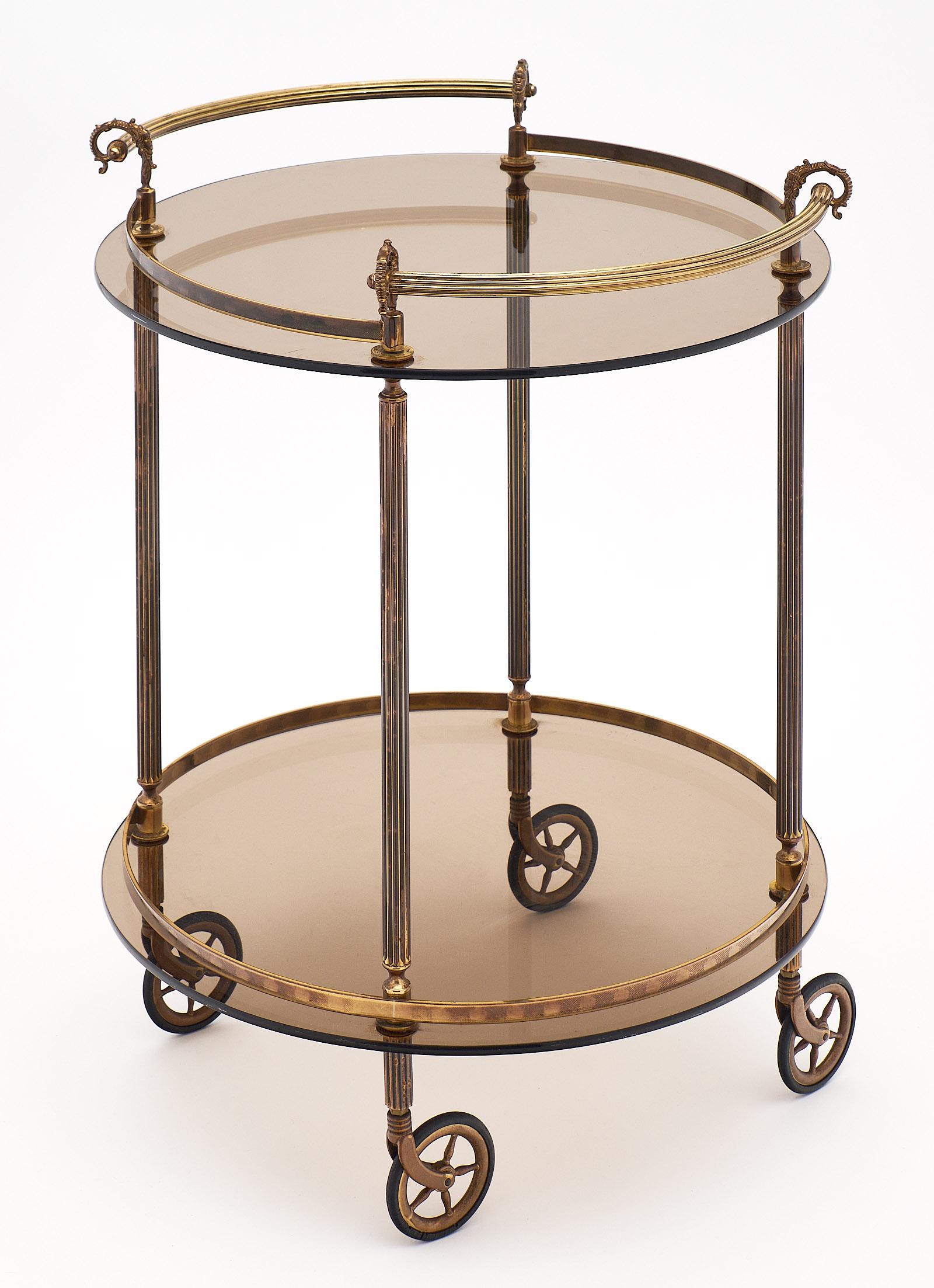 French brass and smoked glass bar cart with two handles and original wheels. This piece has a structure made of brass with curving details. There are two levels of tinted smoked glass. The wheels work very well and are smooth. We love this elegant