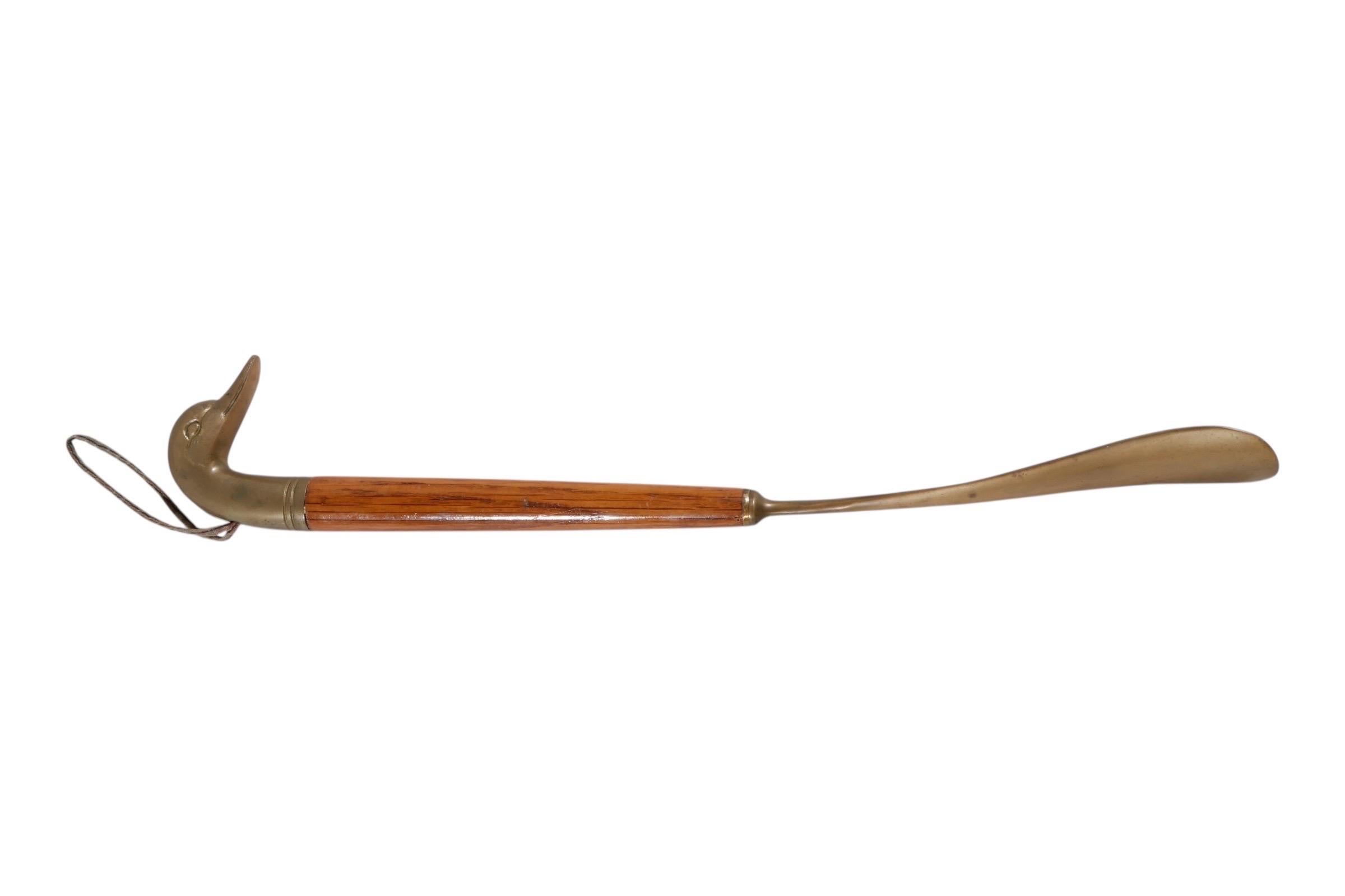 Ein Schuhlöffel aus Messing und Holz. Der Griff ist aus Messing in Form eines Entenkopfes gegossen und mit einer Schnur versehen, um die Aufbewahrung zu erleichtern. Der runde Holzstiel endet in einer traditionellen Schuhlöffelform aus Messing. 