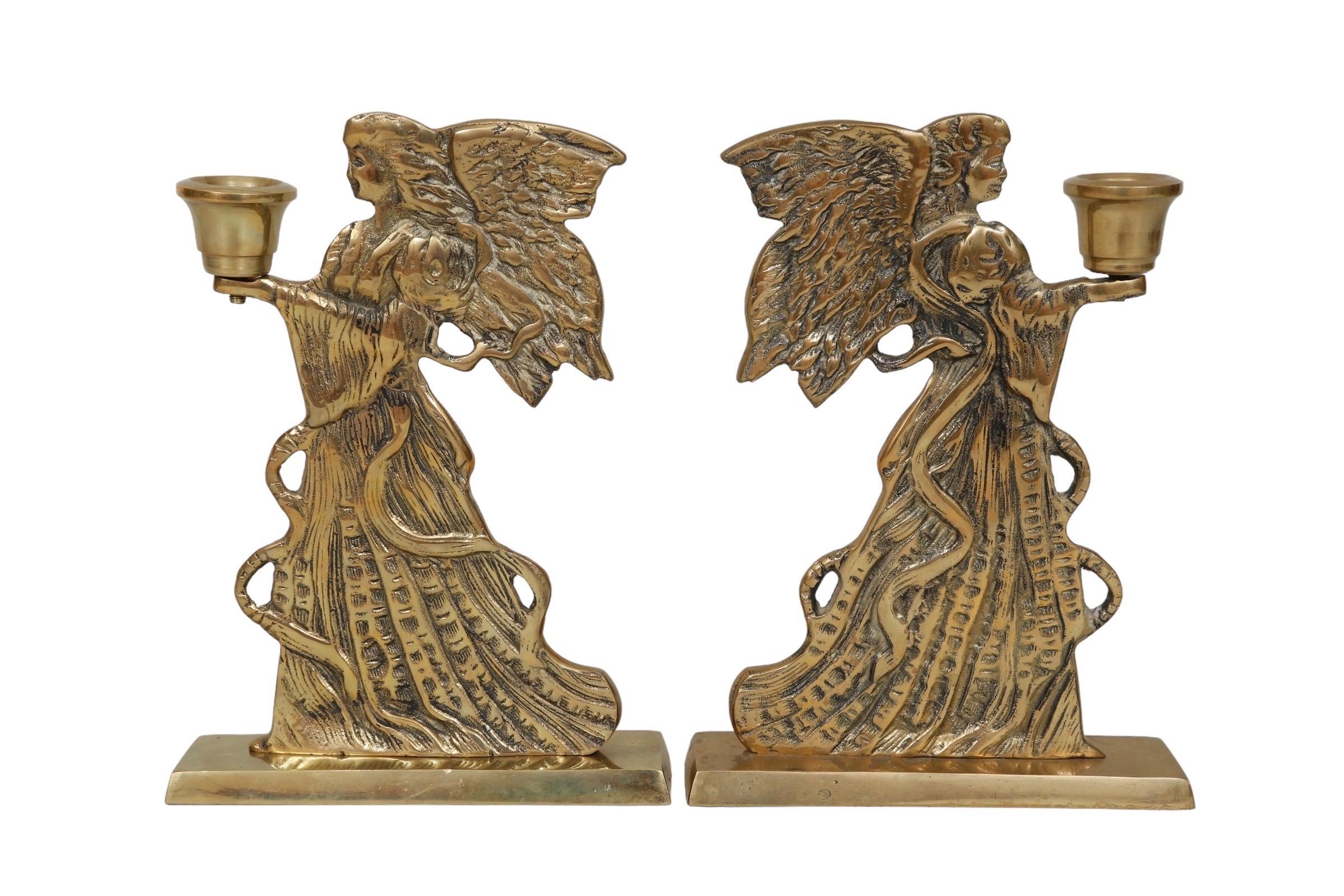 A pair of brass angel candlestick holders. Pressed brass figures are detailed on both sides with feathered wings, flowing gowns and ribbons. Hands extend forward to hold simple brass sconces. Set on rectangular brass bases. Dimensions per candle