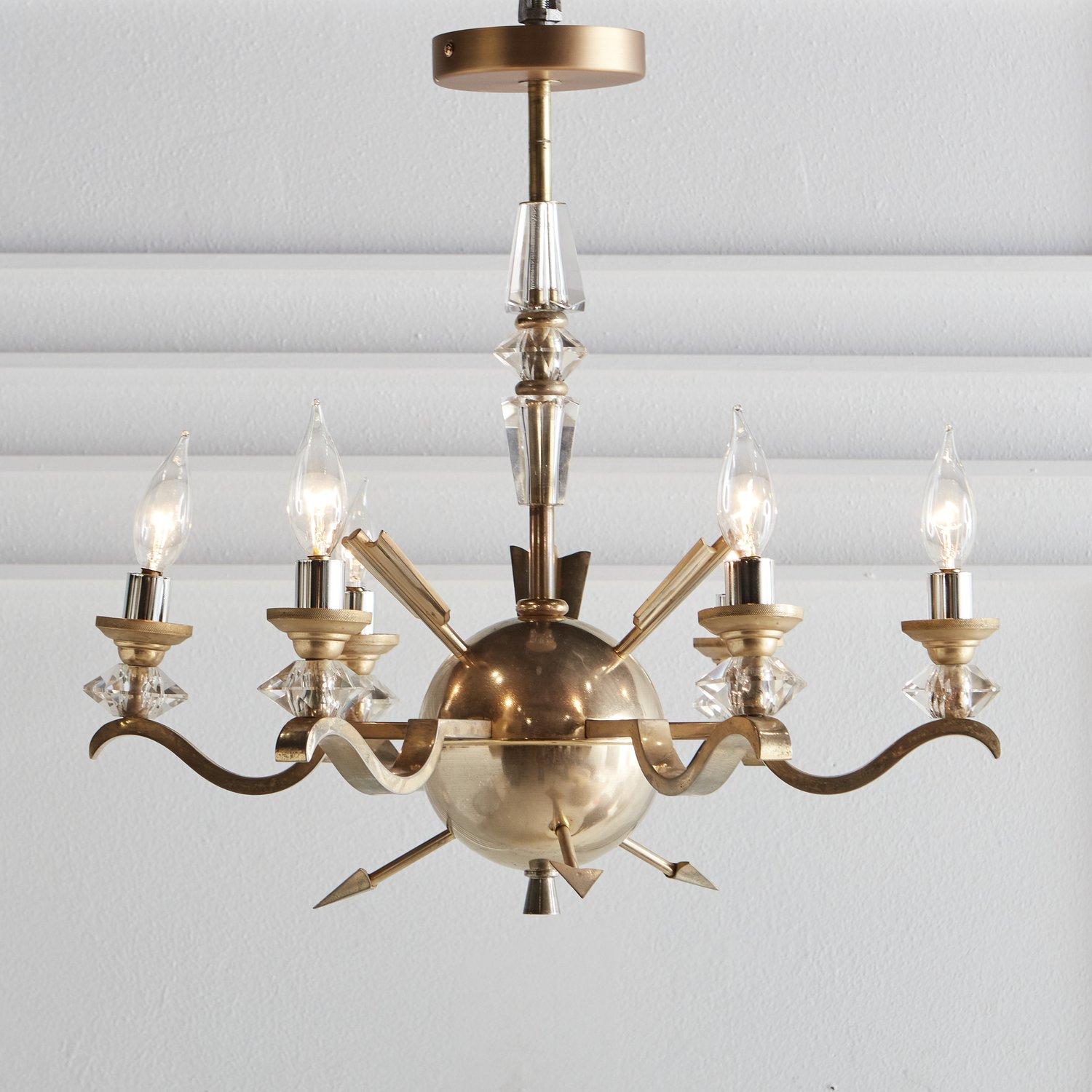 A vintage brass chandelier sourced in France featuring arrow motifs and faceted crystal details. This chandelier has a spherical body with six unique curved arms and chrome candle sleeves with brass bobeches. Early 20th Century.