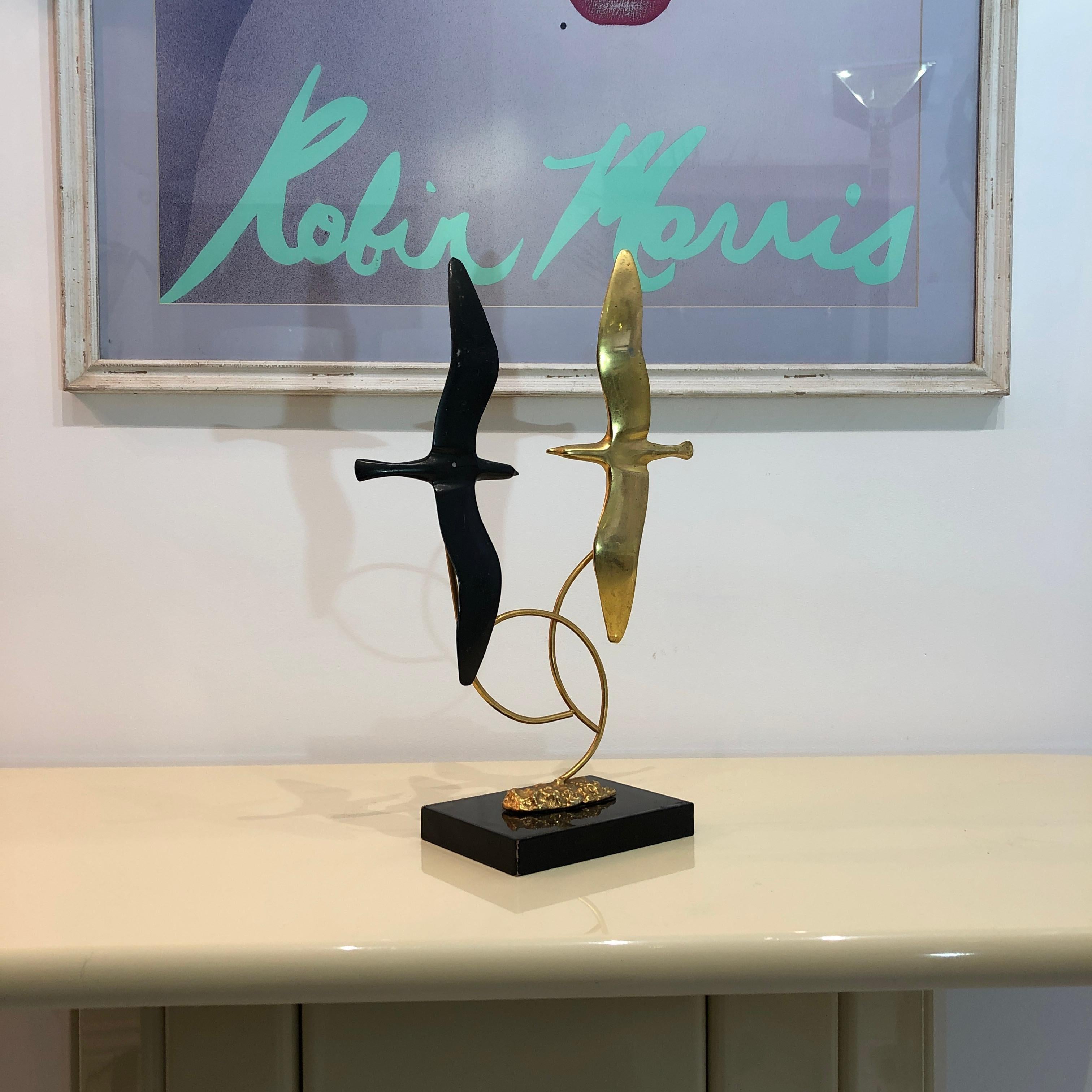 A simple and elegant figurative brass sculpture of two flying birds, one composed of polished brass and one of patinated brass. The swirly brass supports give this piece an undulating and organic kinetic effect of two birds in flight. A black,