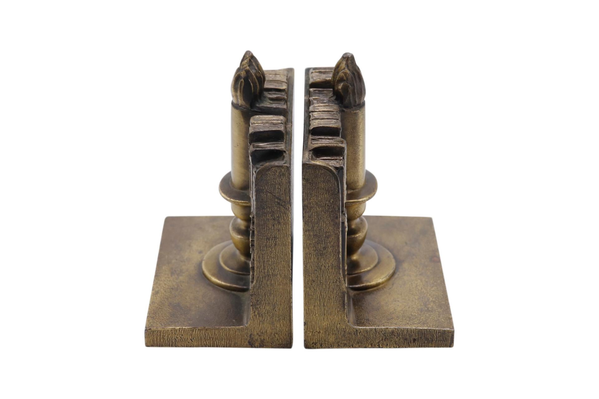 A pair of American made brass bookends cast in the shape of a candle stick in front of bookshelves. Underneath is finished with red felt. Makers label reads “A product of the finest American Craftsmanship. Philadelphia MFG Co. Philadelphia, PA.