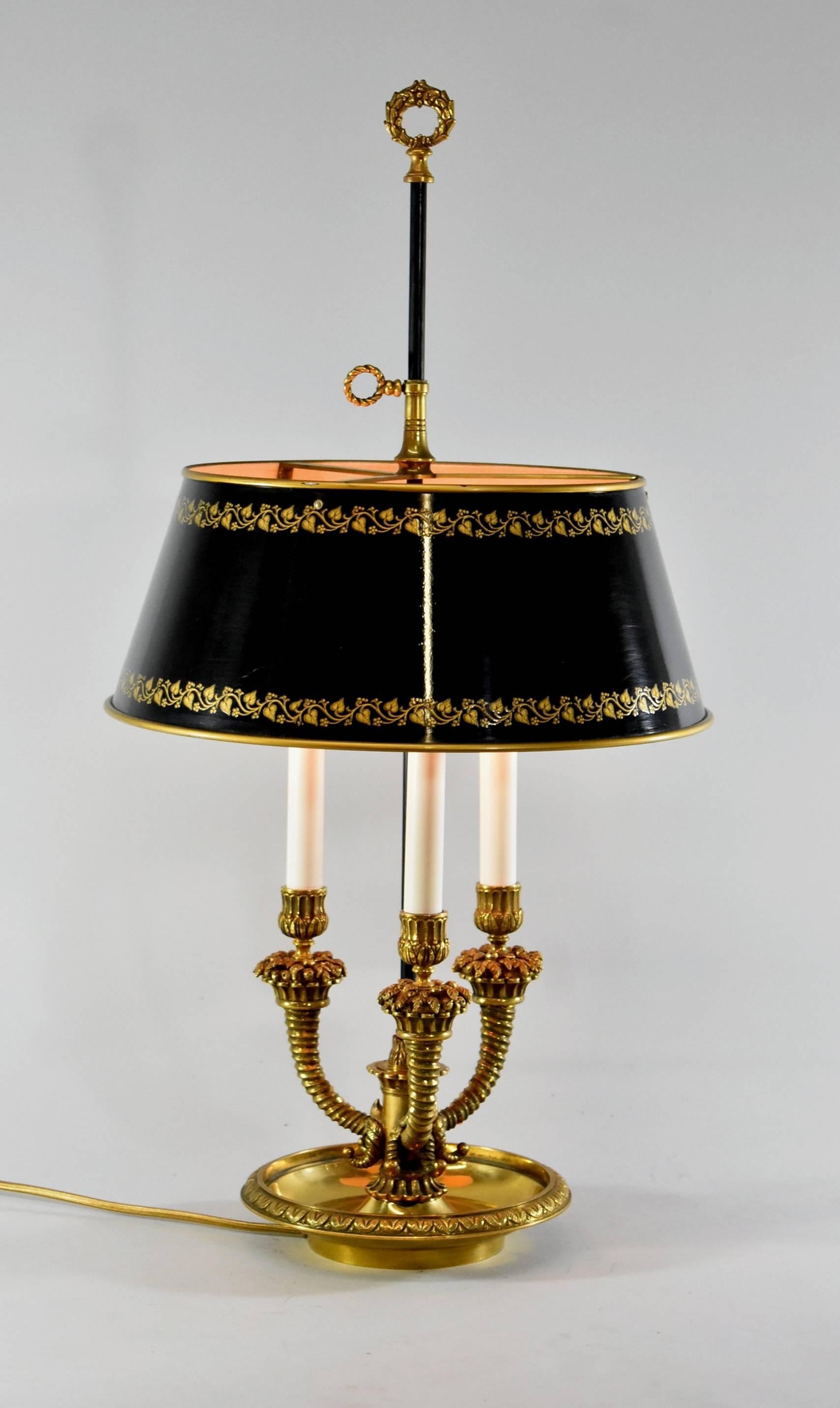 An elegant French style brass Bouillotte lamp. Fitted with three cornucopia form lights with candelabra size sockets. It features a black metal adjustable shade with gold leaf detail and a wreath form finial. It would be a distinctive addition to a