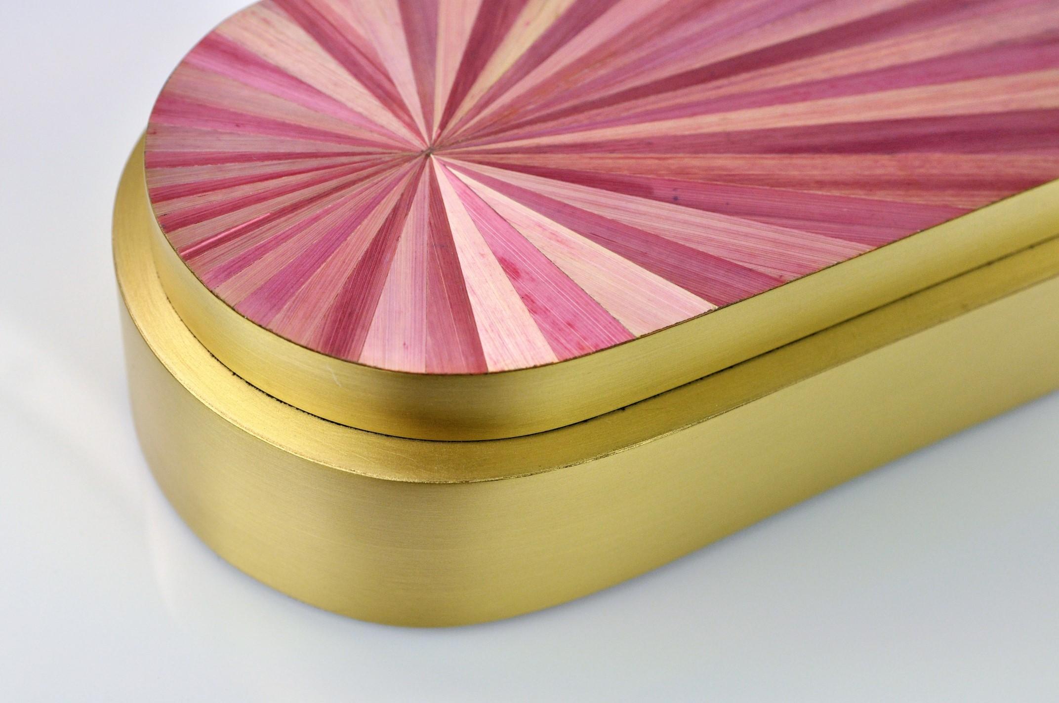 Cette jolie boîte en laiton est en laiton brossé avec un couvercle recouvert de marqueterie de paille. 
Le design radial de la paille rappelle la grande période Art déco tandis que l'élégante couleur rose contraste parfaitement avec le laiton