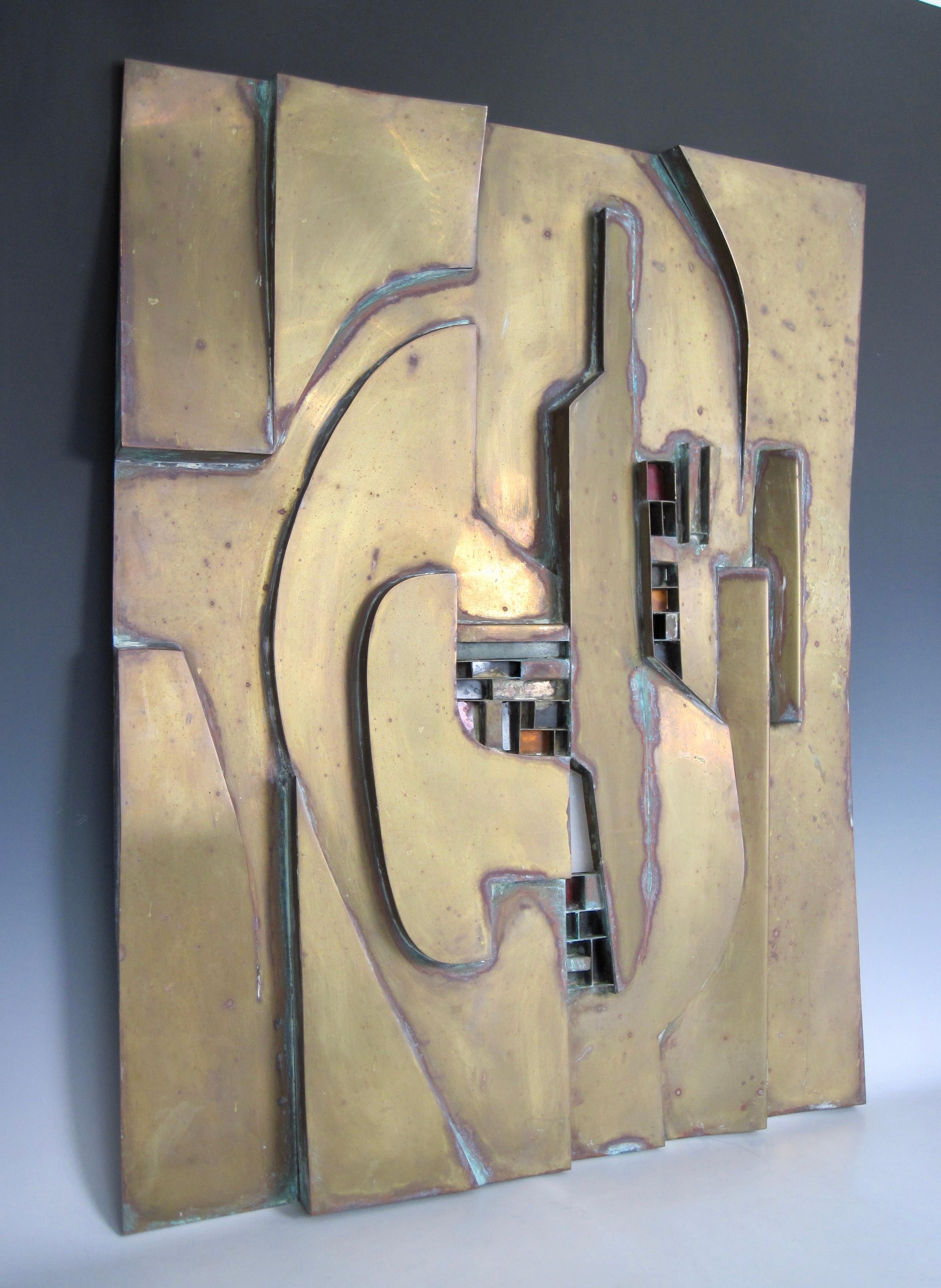 A brutalist brass sculpture of varying advanced and recessed geometric shapes. Fitted stained glass pieces in some smaller square and rectangular pockets. 
We love the geometric shapes with the brutalist architectural feel.
Wire hanger attached on