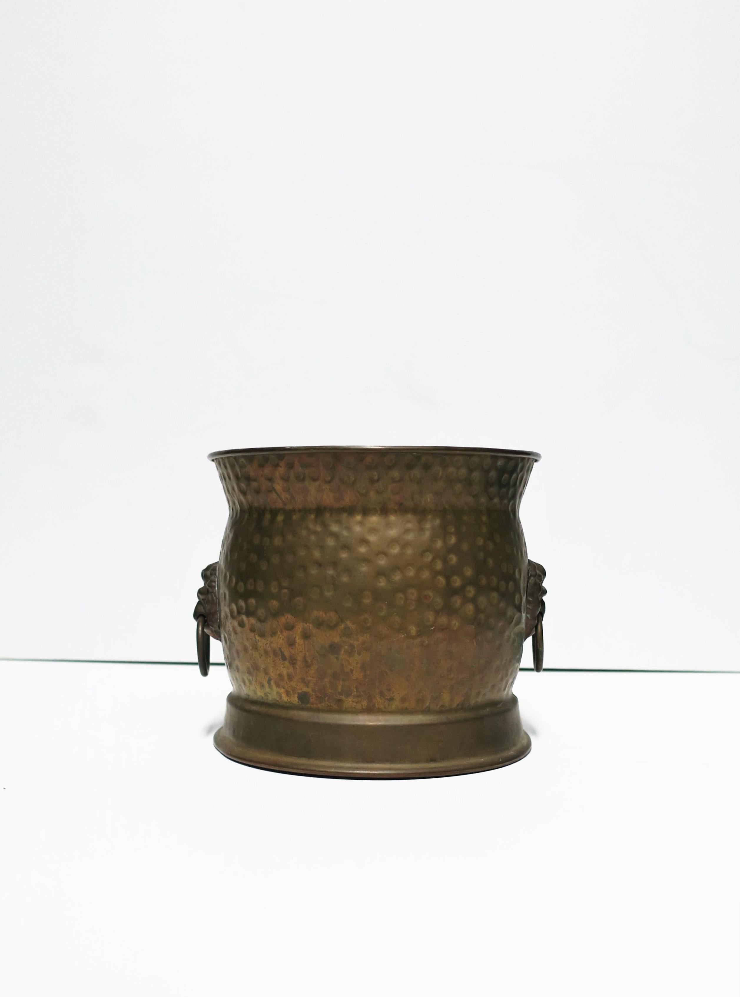 An English antique bass cachepot (plant or flower holder) with hammered design and lion head and ring detail on sides, in the Regency style, early 20th century, England. Piece is unpolished with beautiful age patina to brass. Dimensions: 7”D x 7.38”