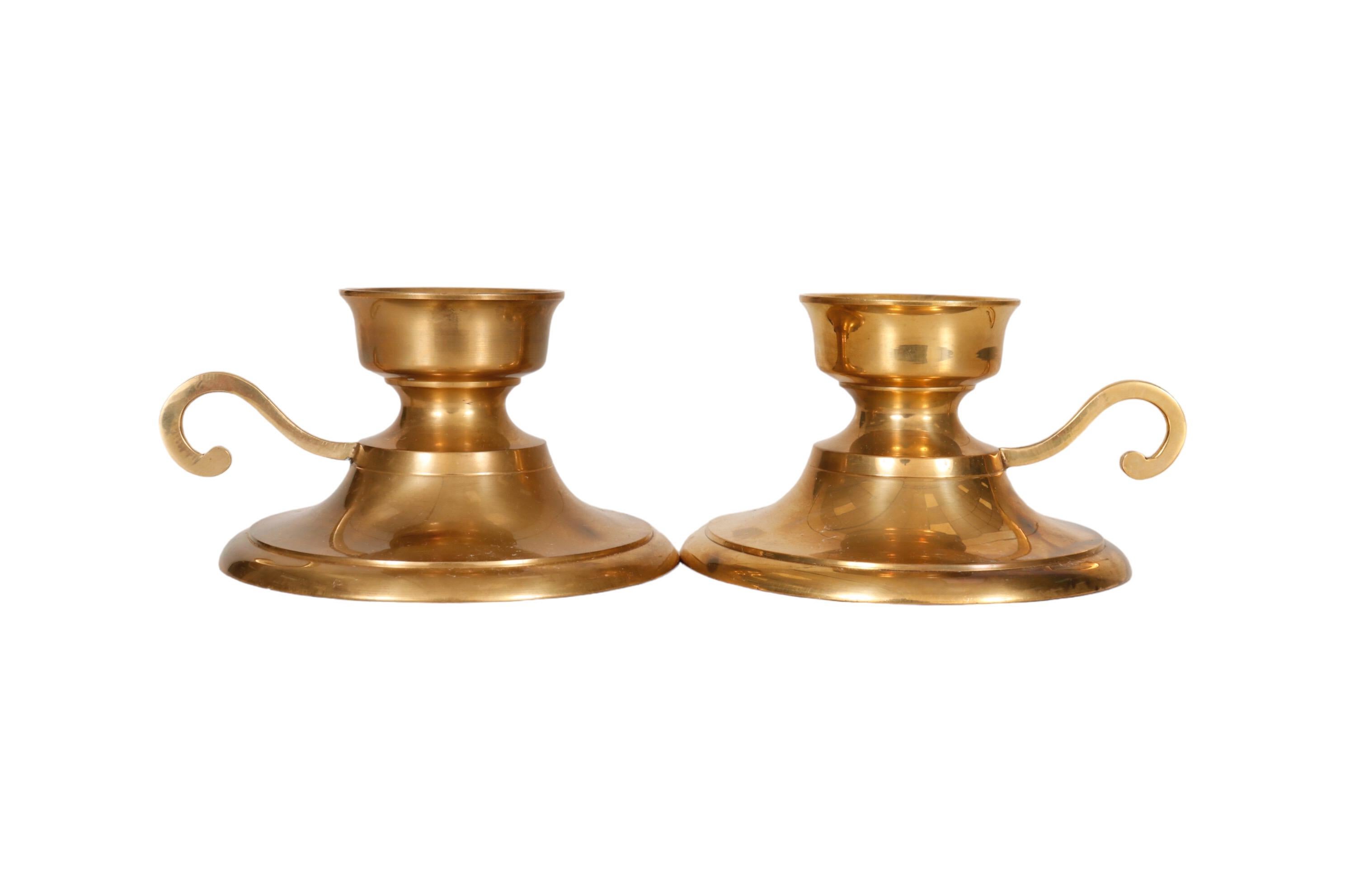 A pair of brass chamber stick candle holders. Each has a short capital with a spike to secure the candle, with a curled brass handle above a wide base. Dimensions per candle holder.