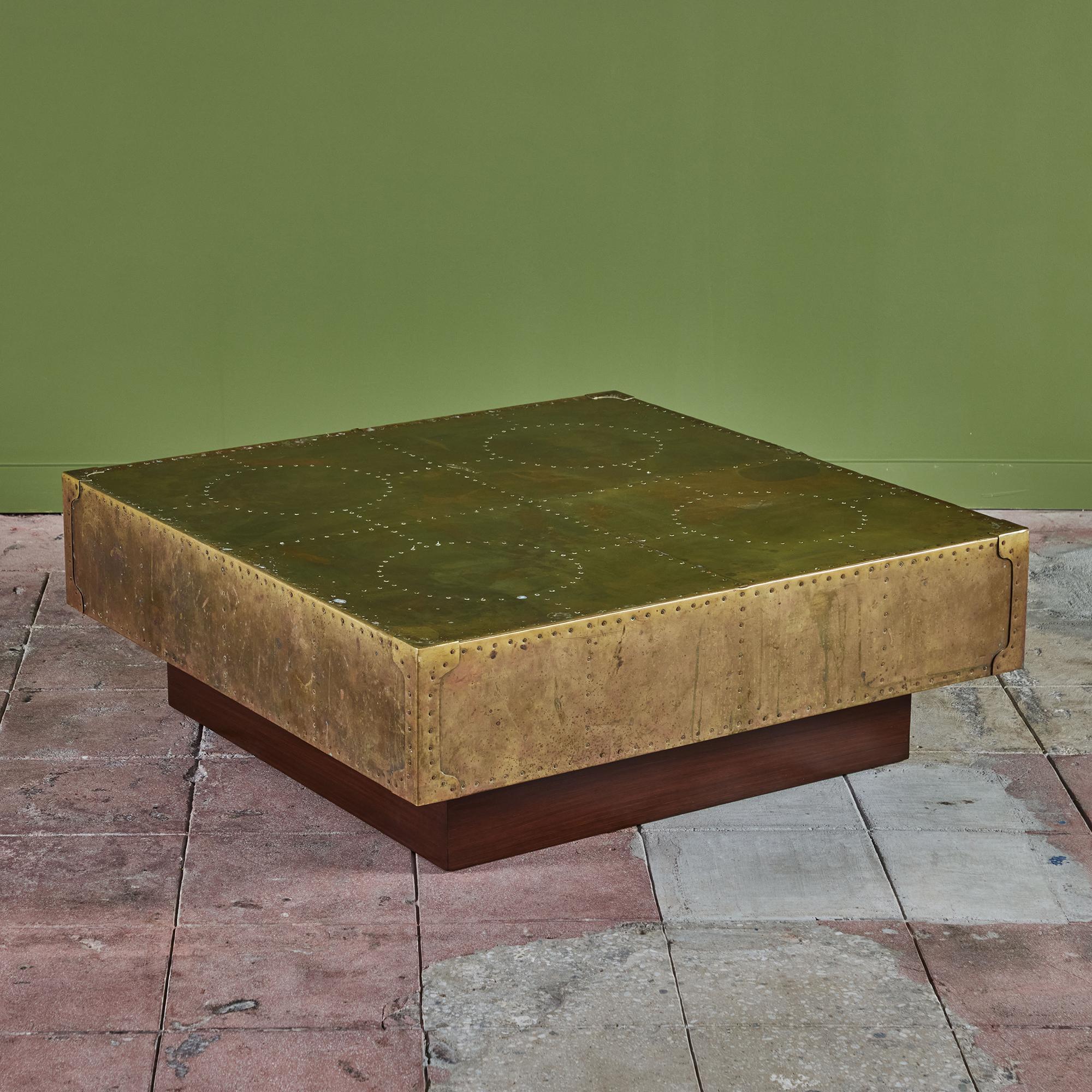 Square brass clad coffee table on a walnut plinth base in the style of Sarreid. The table has a brass riveted detailing. The table top features a geometric pattern of squares and circles showcased by the rivets. The brass sheets that make up this