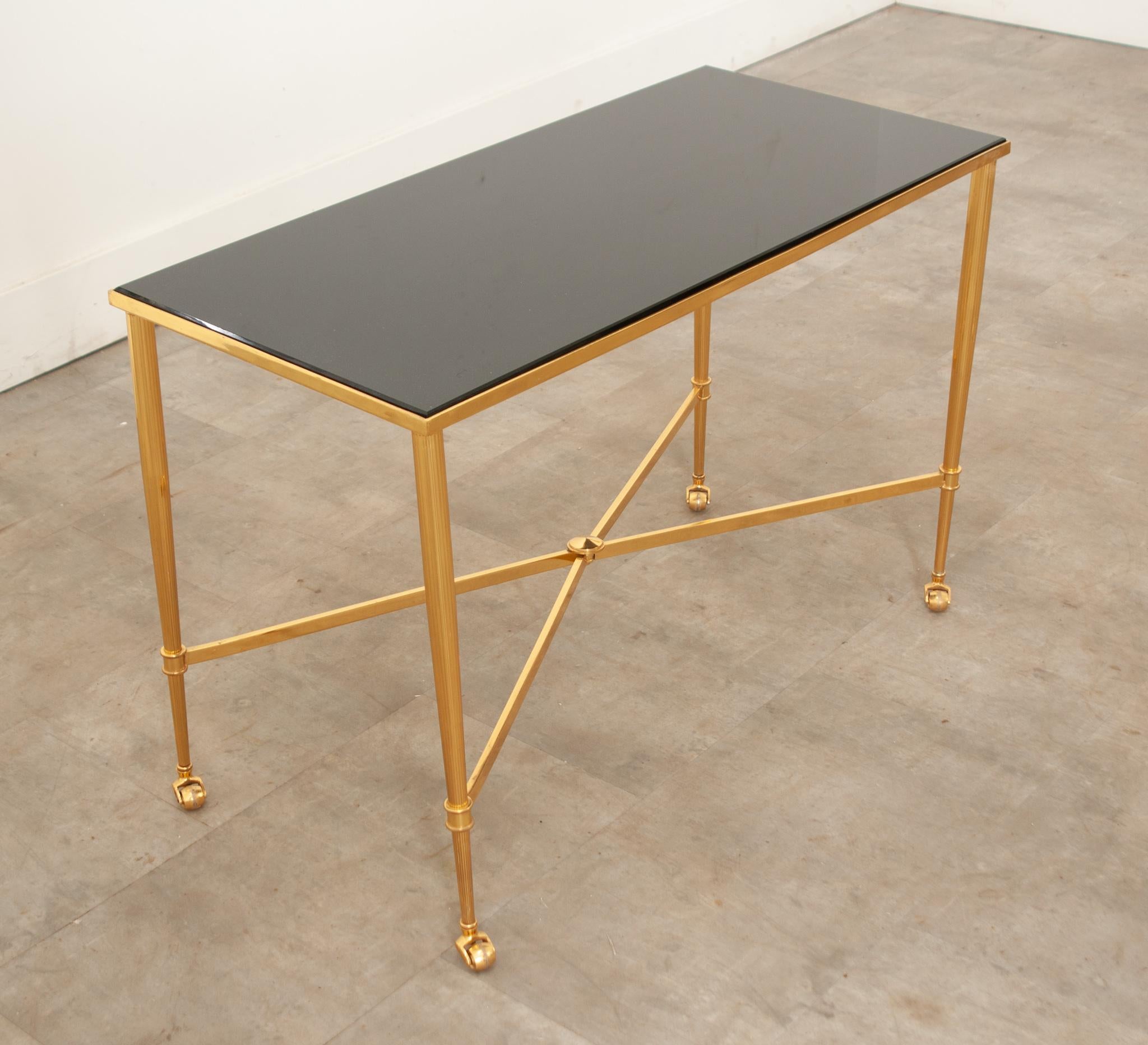 A sophisticated French brass cocktail table in the style of Masion Jansen. Topped with a removable inset black stone over tapered legs connected to an X-shaped stretcher with smooth rolling casters. Vibrant and stunning, this table will add class to
