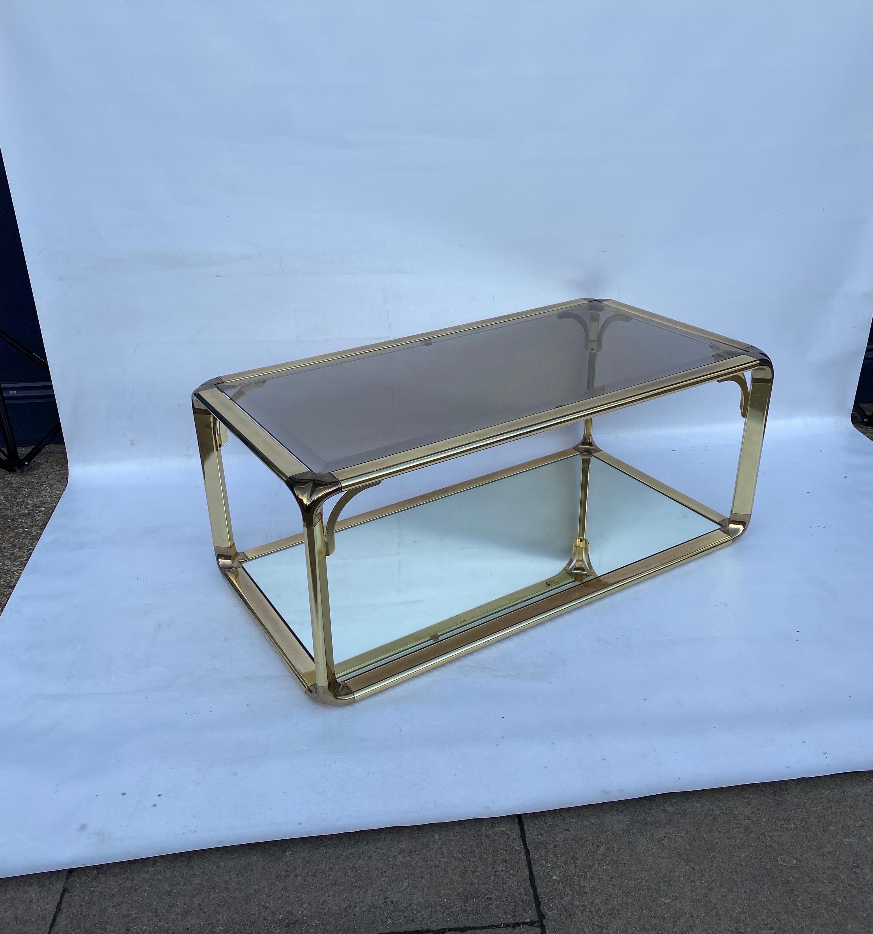 Une table basse agréable avec des bords, des coins et des lignes courbés.
Cadre en laiton avec joints d'angle détaillés, avec un verre biseauté légèrement fumé sur le dessus et un miroir transparent sur le dessous. 
Une pièce facile à utiliser et à