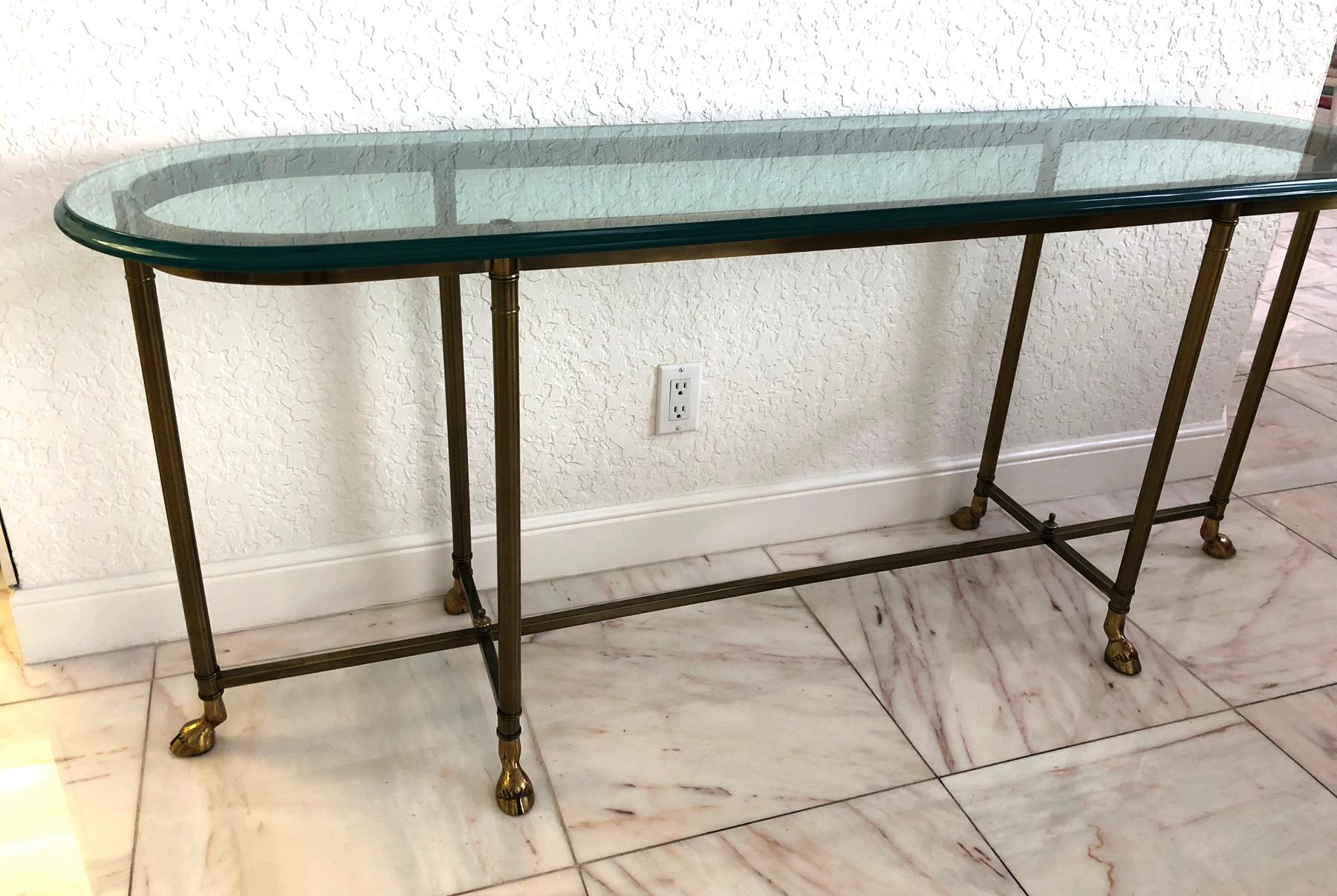 Large Hollywood Regency brass console table with thick beveled edge glass top. Large hoof feet in the style of Jansen. Rounded ends add a soft look to table. Measures: 65 wide by 18 deep by 28 inches high.