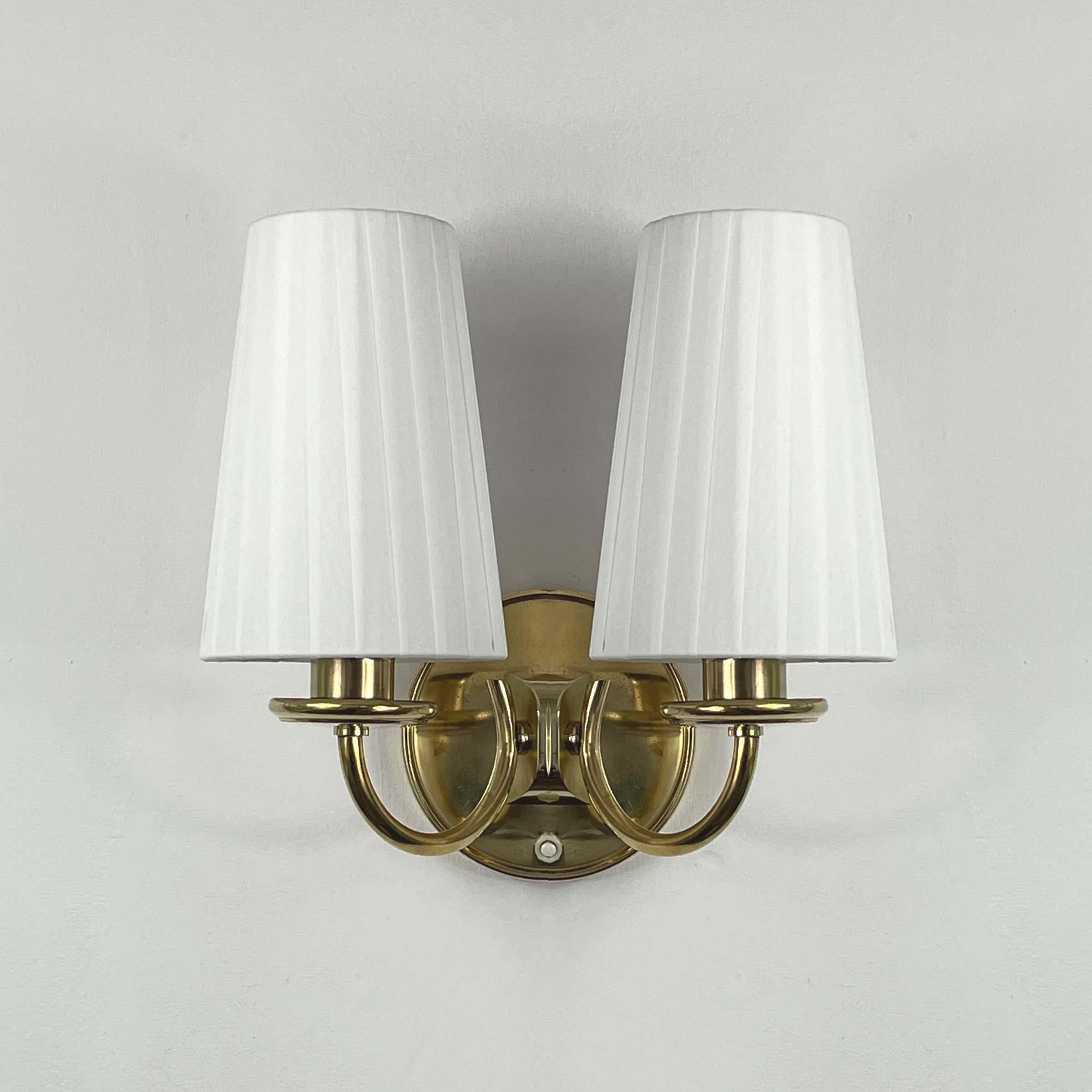 Questa lampada da parete in stile Art Déco scandinavo è stata progettata e prodotta in Svezia negli anni '40.

La lampada ha un design classico ed elegante che combina ottone e tessuto. Presenta due bracci luminosi, ognuno dei quali sostiene un