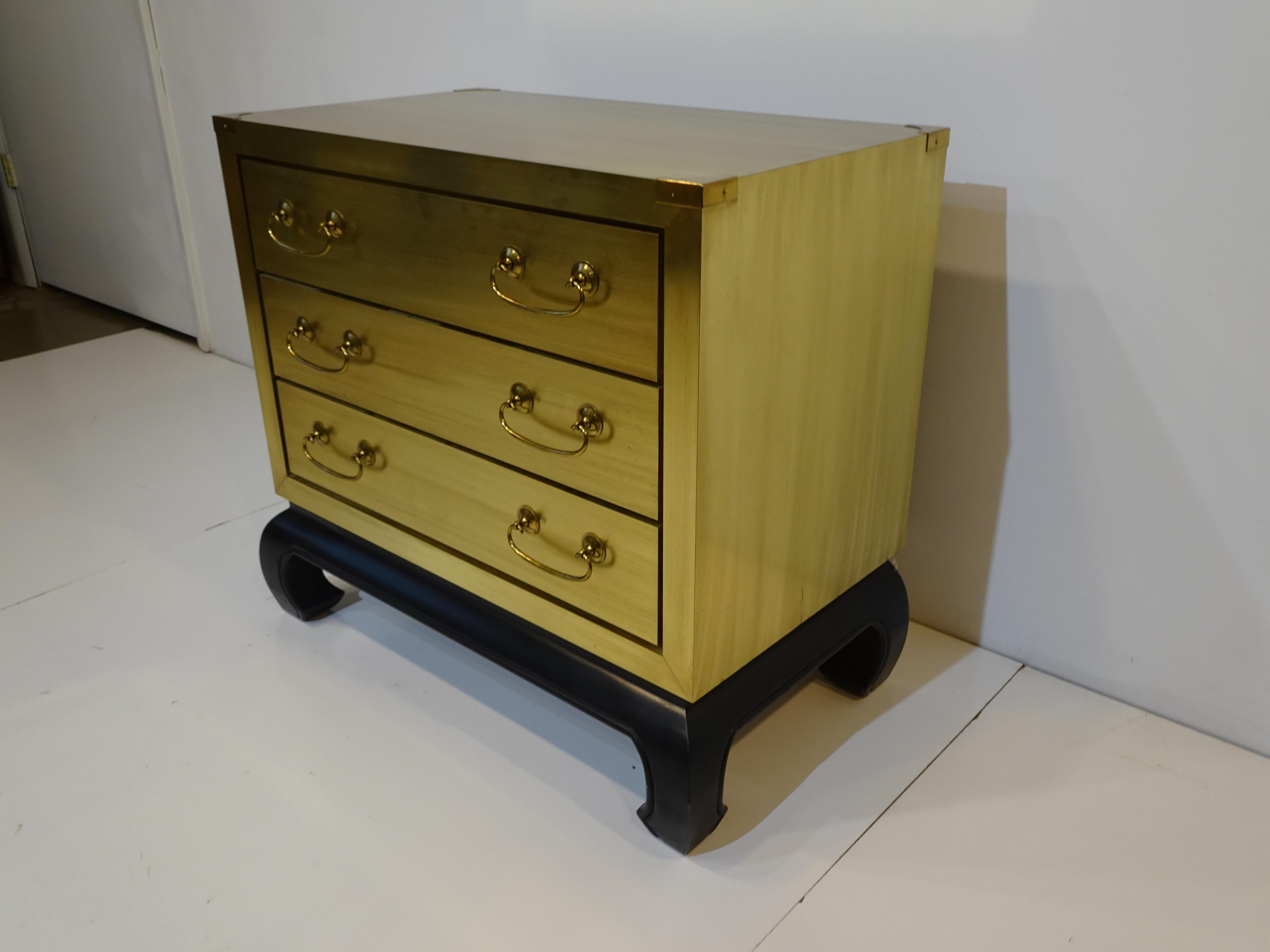 A three drawer small dresser chest with brass and wood construction having a satin black decorative base and large brass pulls. Very well crafted with wood drawers and corner details to the dresser in brass, a piece that would work in any interior,