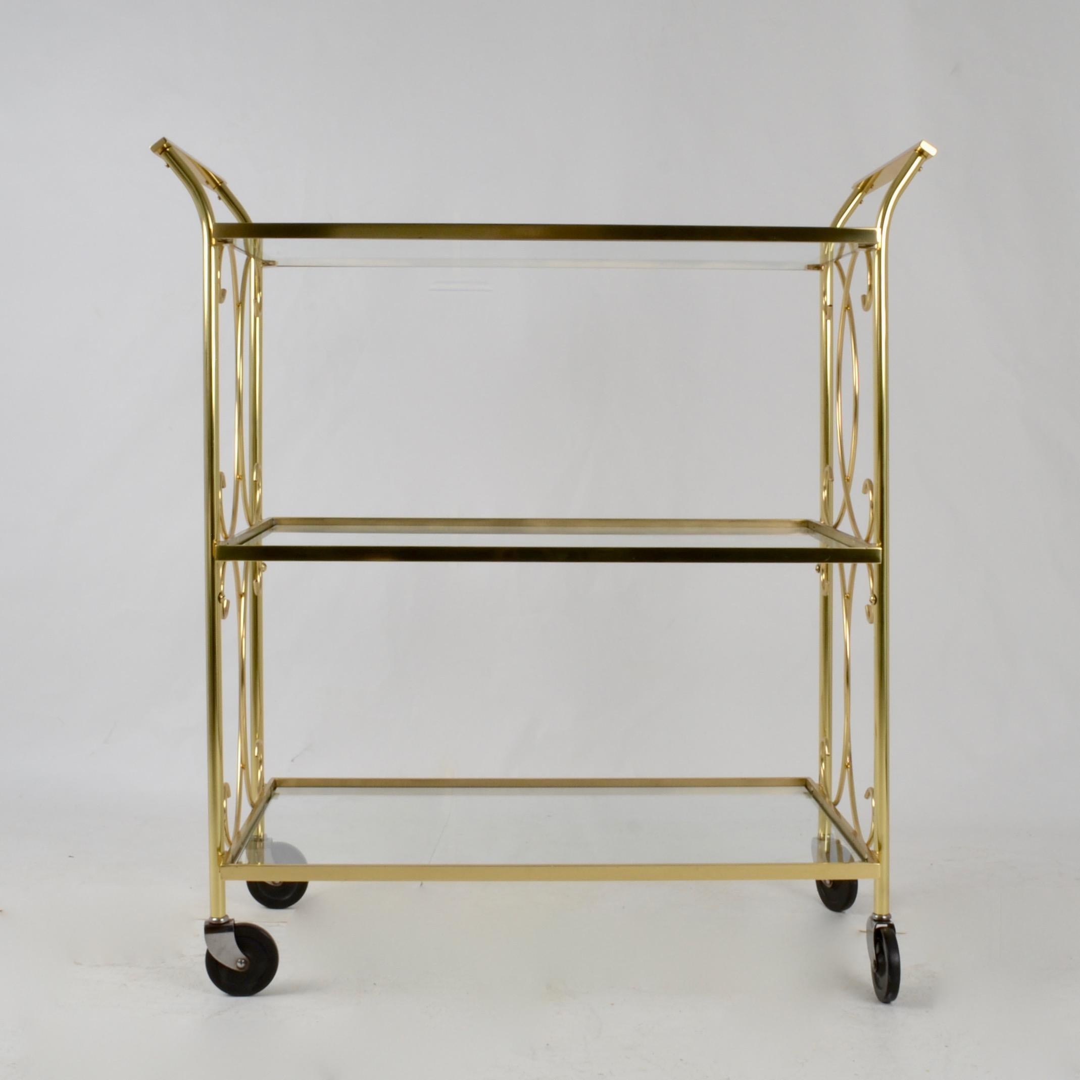 Charming solid brass cocktail cart with three levels and whimsical curved decorations. Black composite wheels. Clear glass shelves. Brass has been polished and lacquered.