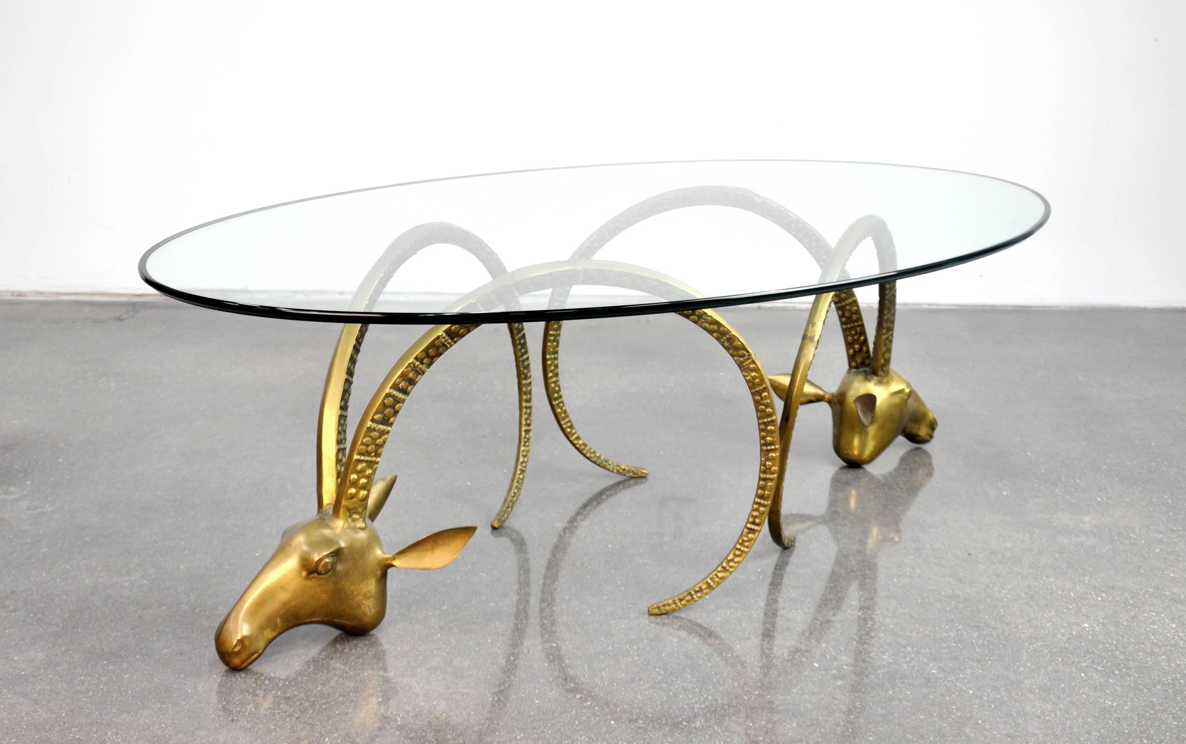 A striking Mid-Century Modern ram's head cocktail table in the style of Alain Chervet, dating from the 1970s. The Hollywood Regency table features an oval bevelled glass top resting on a pair of brass ibex heads. The bases can also be used
