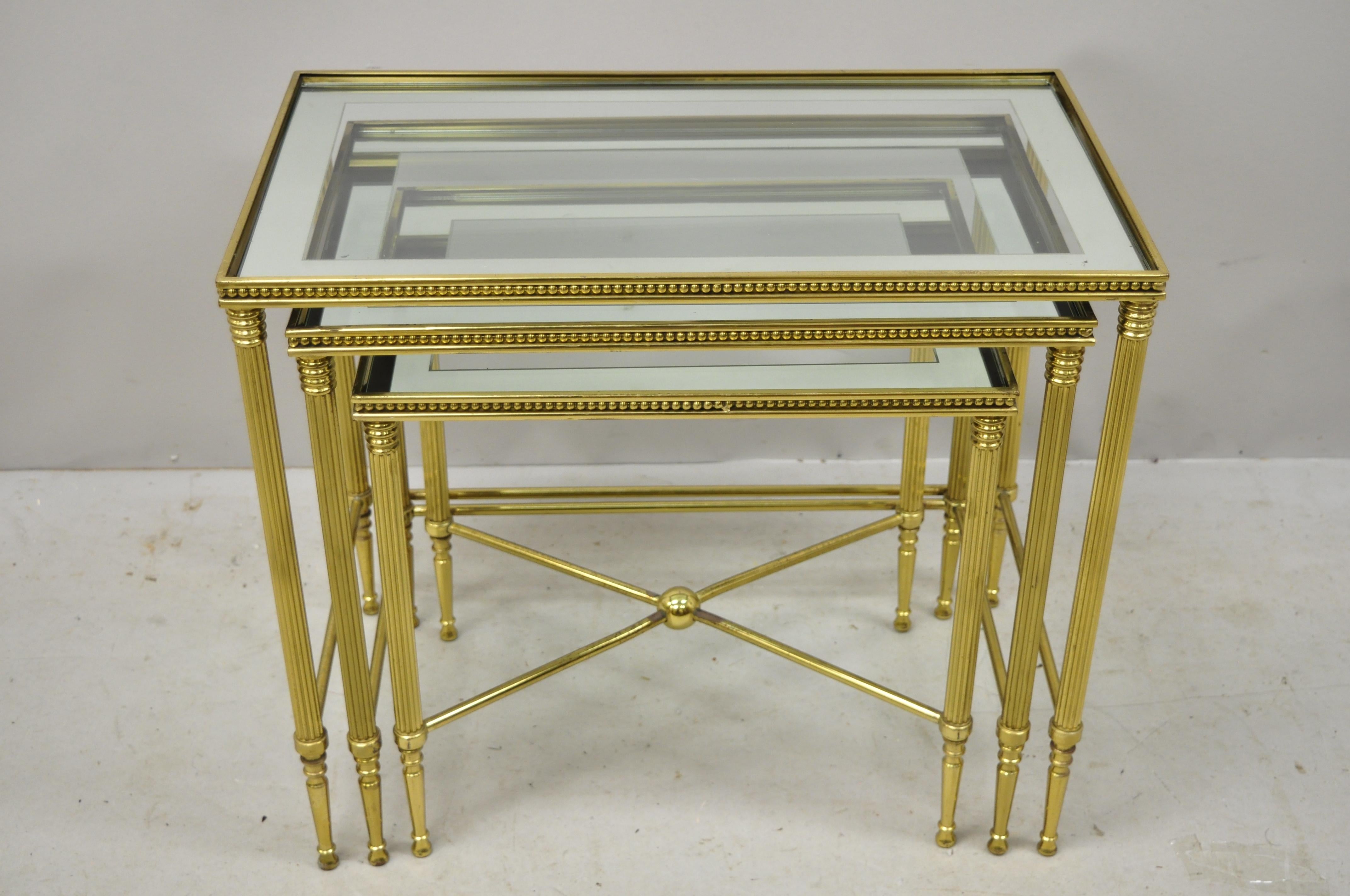 3 brass Italian glass top Hollywood Regency side nesting tables with tapered legs. Set features glass tops with mirror borders, brass beaded trim, ball form stretcher, brass construction, sleek tapered legs, quality Italian craftsmanship, circa