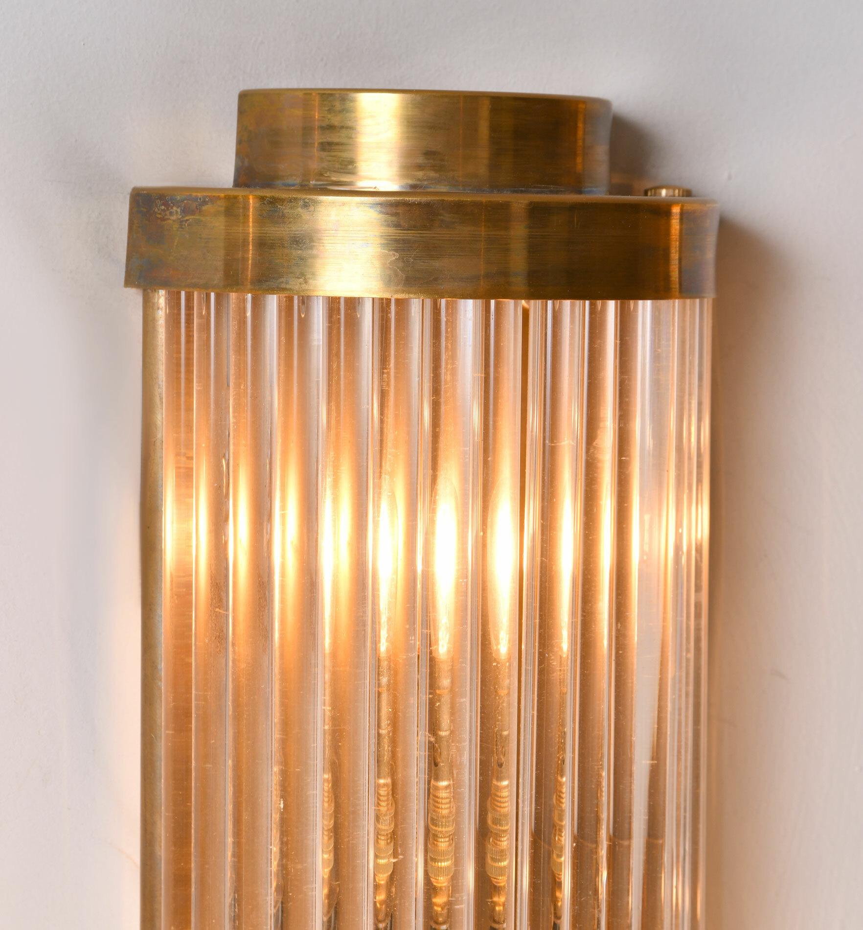 This ‘Ravello’ wall light takes its inspiration from a timelessly classic Art Deco design.
Multiple Italian glass rods form a semi-circle which is capped, top and bottom by 2 tiers of curved brass. The brass back plate holds two light bulbs and