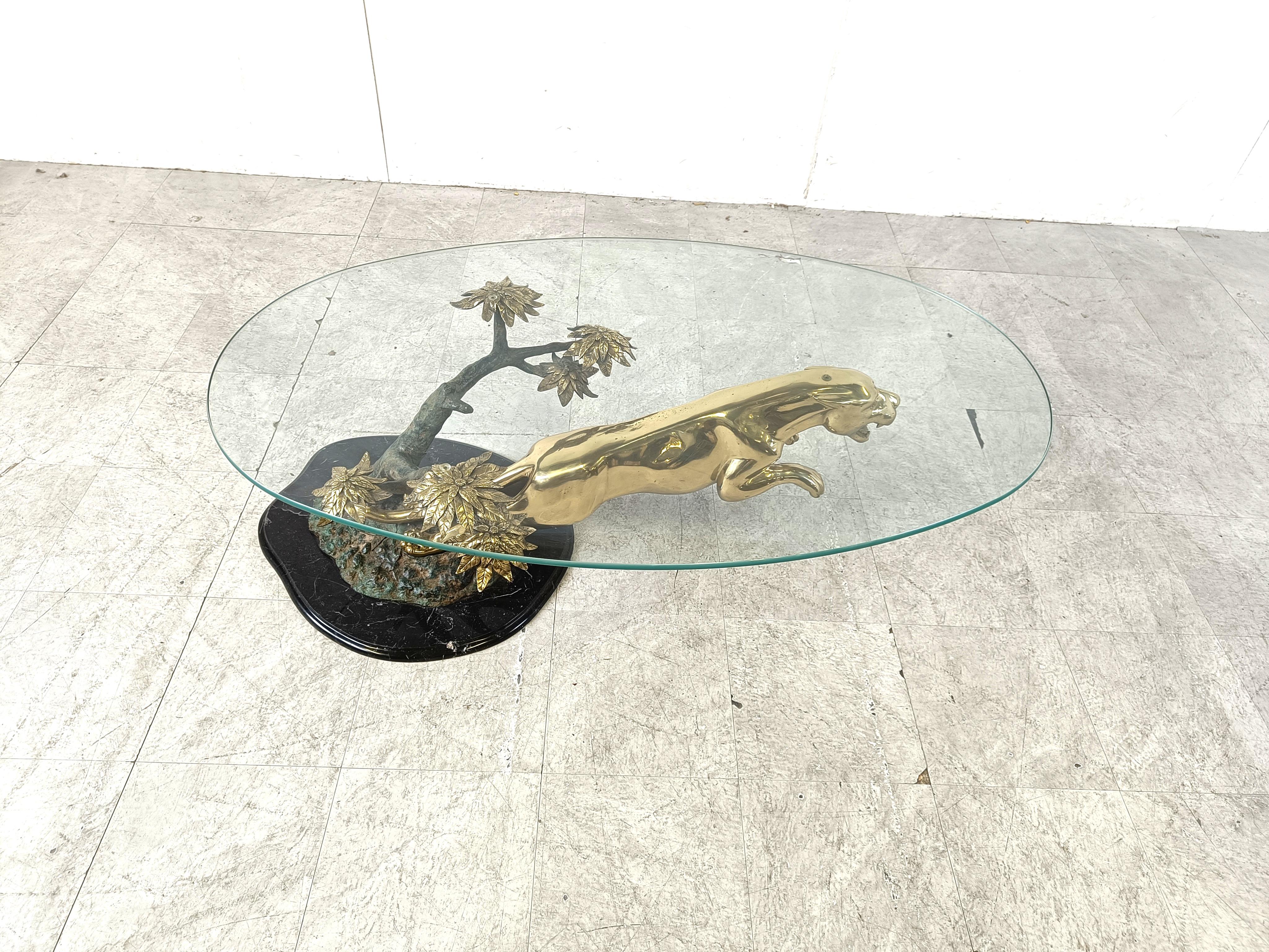 Spectaculaire table basse représentant un jaguar bondissant, joliment sculpté et réalisé en laiton.

Il est monté sur une base en marbre noir qui présente un arbre en bronze avec des fleurs en laiton sculptées, supportant un plateau en verre