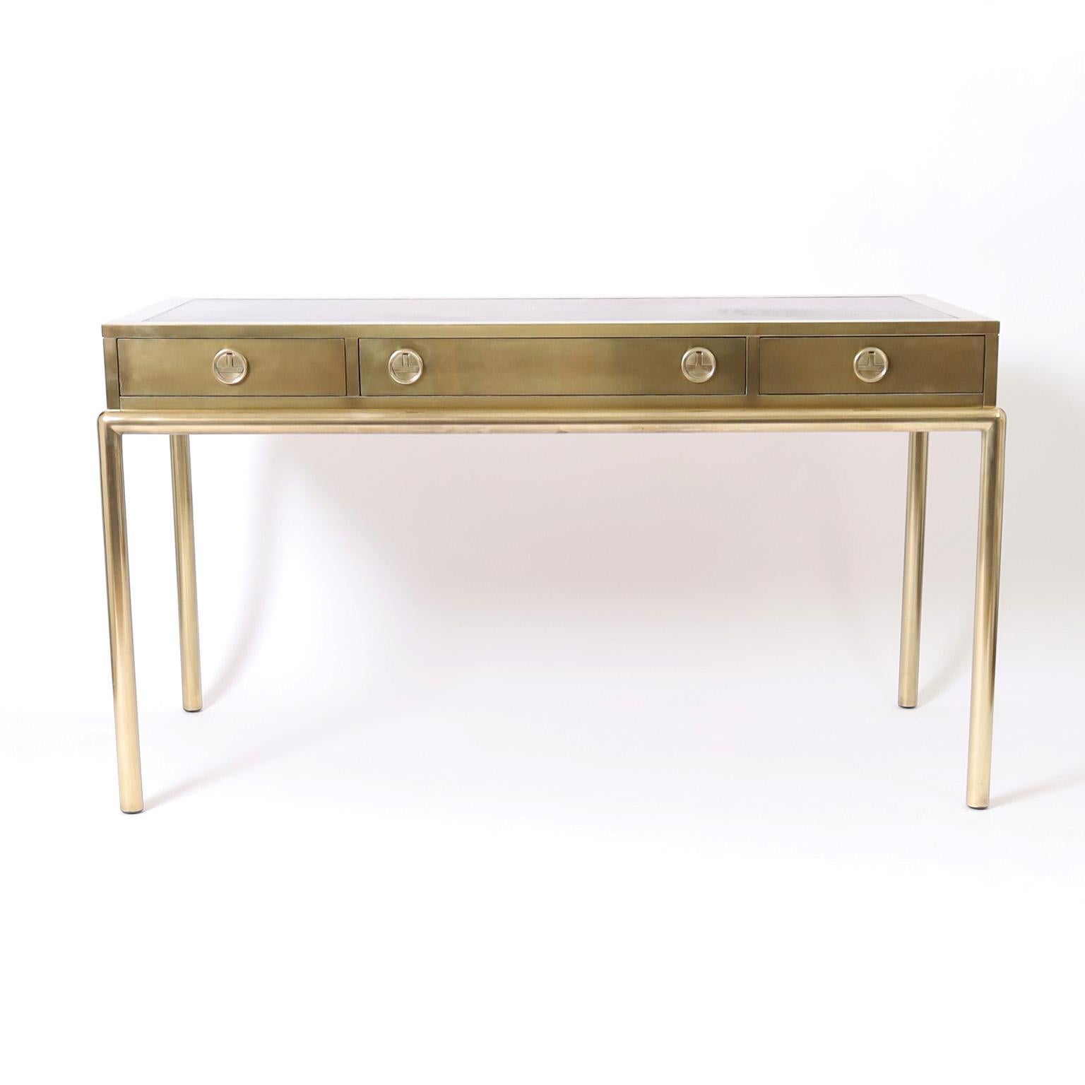 Mid century Mastercraft three drawer desk or writing table crafted in brass with an acid washed finish featuring a tooled leather top, stylized campaign drawer pulls, and a sleek modern design.