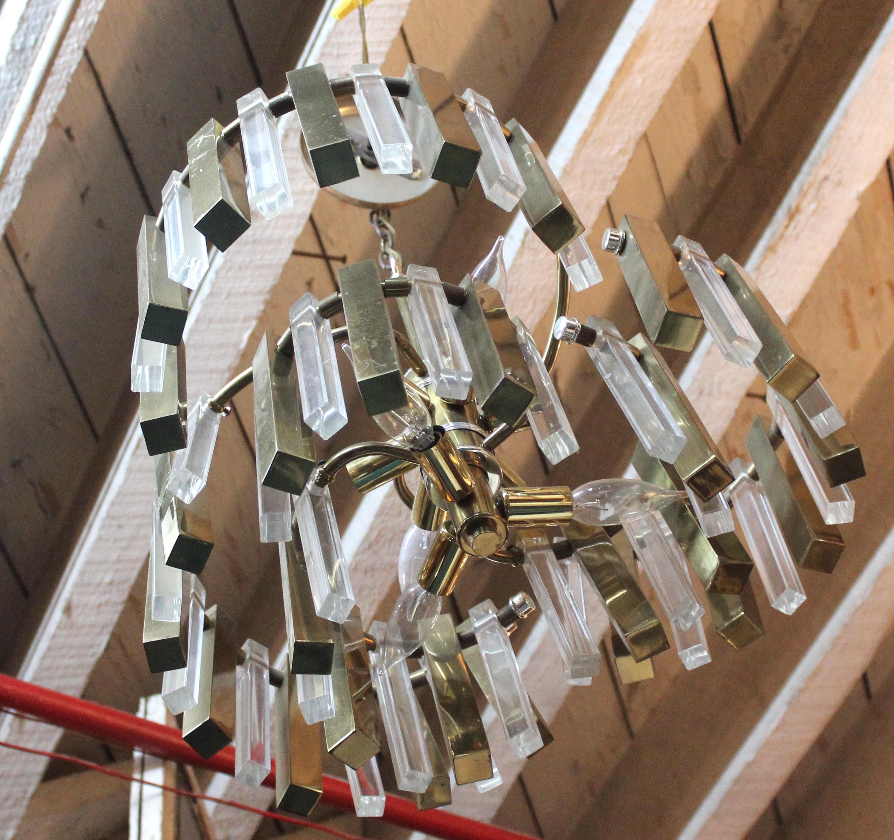 Questo lampadario ha tre bracci vorticosi che sostengono barre alternate di ottone e lucite trasparente che ombreggiano le sei lampadine che circondano la lampada. 
Si prega di confermare la posizione NY o NJ