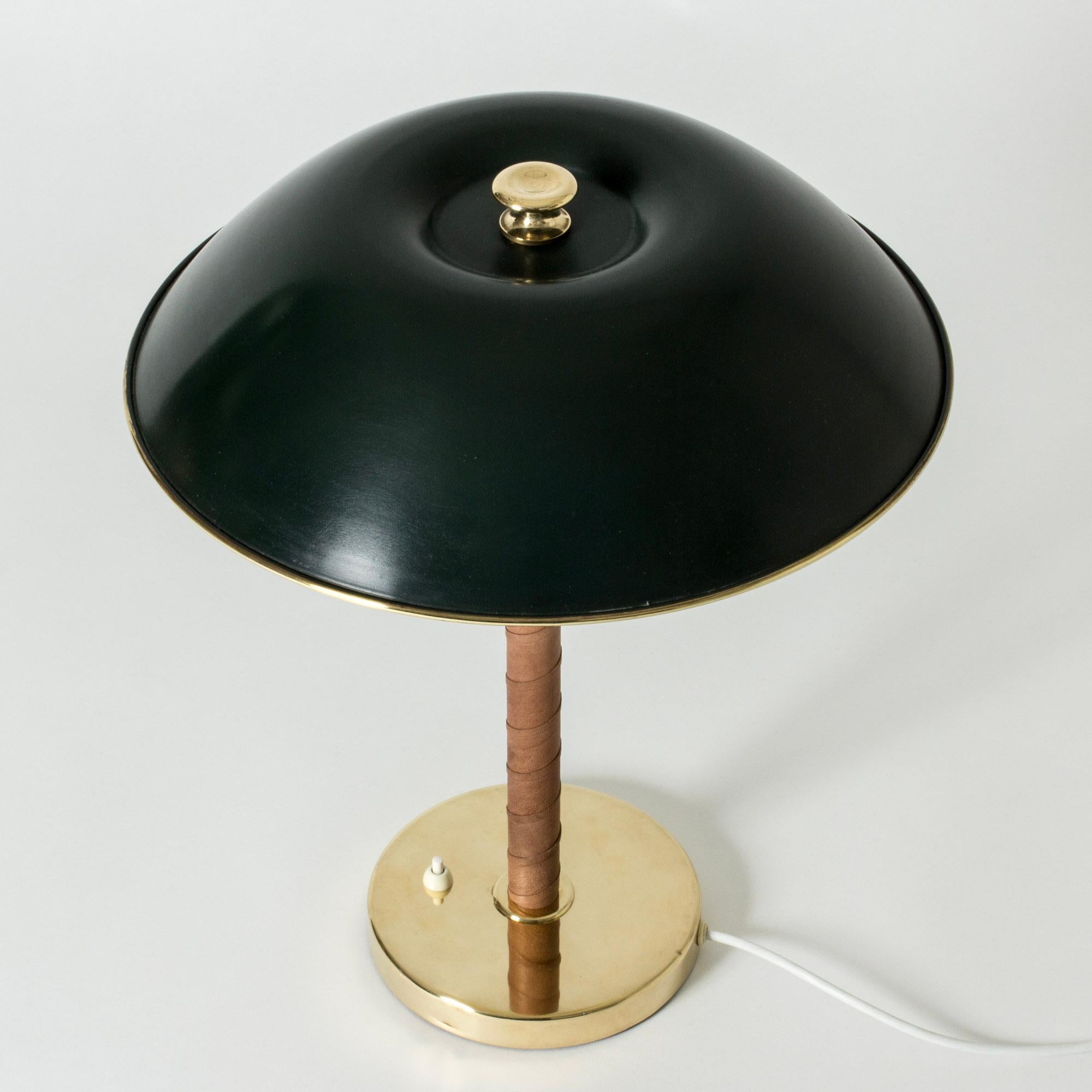 Elegante lampe de table de Böhlmarks, avec une base en laiton, une poignée enroulée en cuir et un abat-jour en métal laqué vert orné d'un bord et d'un bouton en laiton.