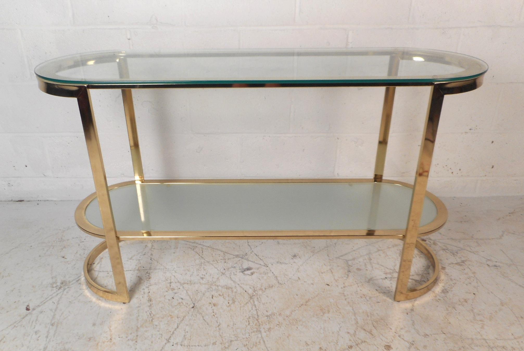 This beautiful vintage modern coffee table features a unique oval shape with two tiers of thick glass shelves. A stylish design with a flat bar brass frame and a lower frosted glass tier adding to the allure. This stylish piece looks great behind