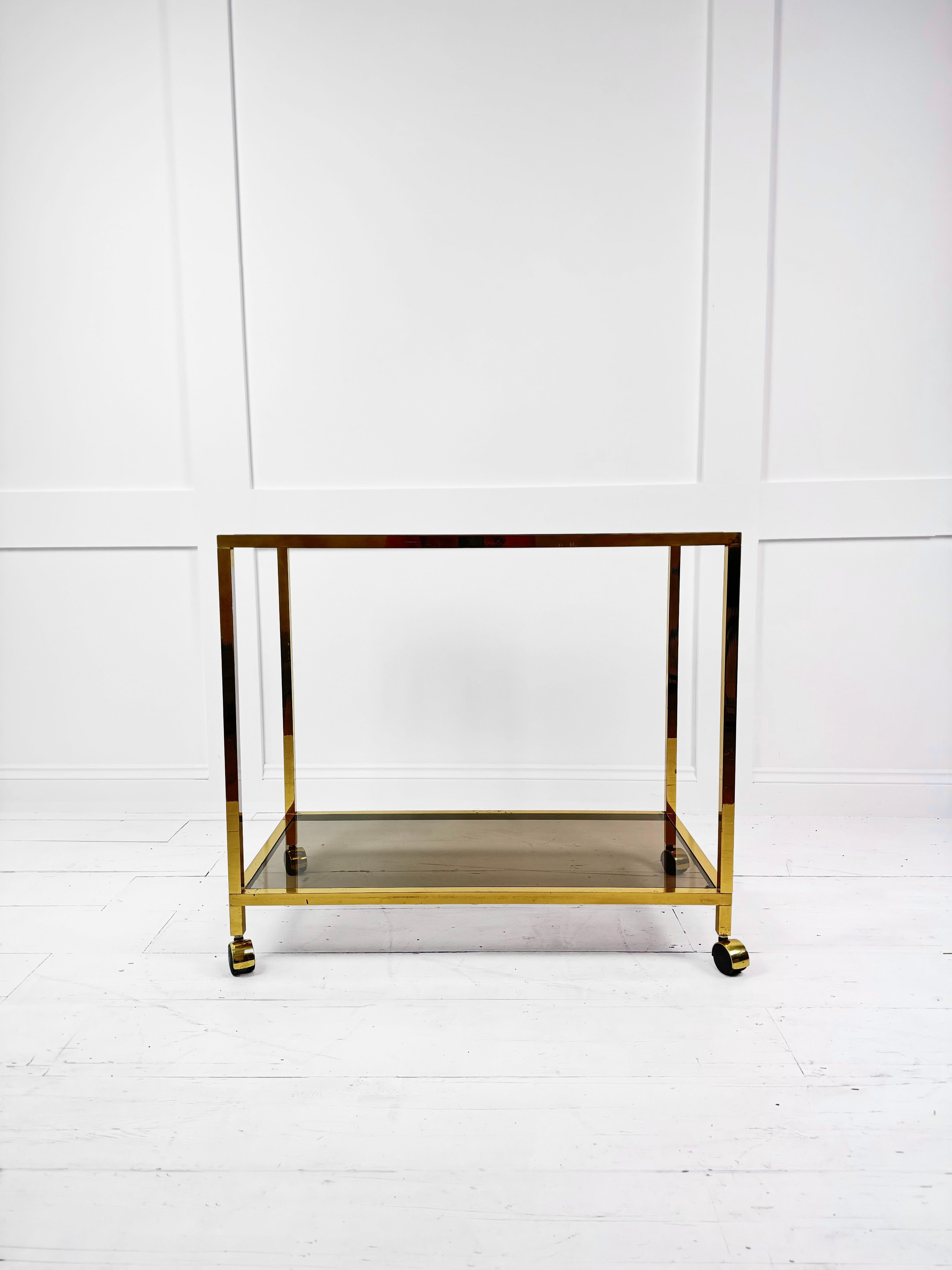 This vintage Brass Midcentury Side Table / Drinks Trolley from Belgium is a stunning piece of furniture from the 1970s. Crafted in Brass and featuring a smoked glass top and lower shelf, this table exudes a timeless Midcentury aesthetic. Its