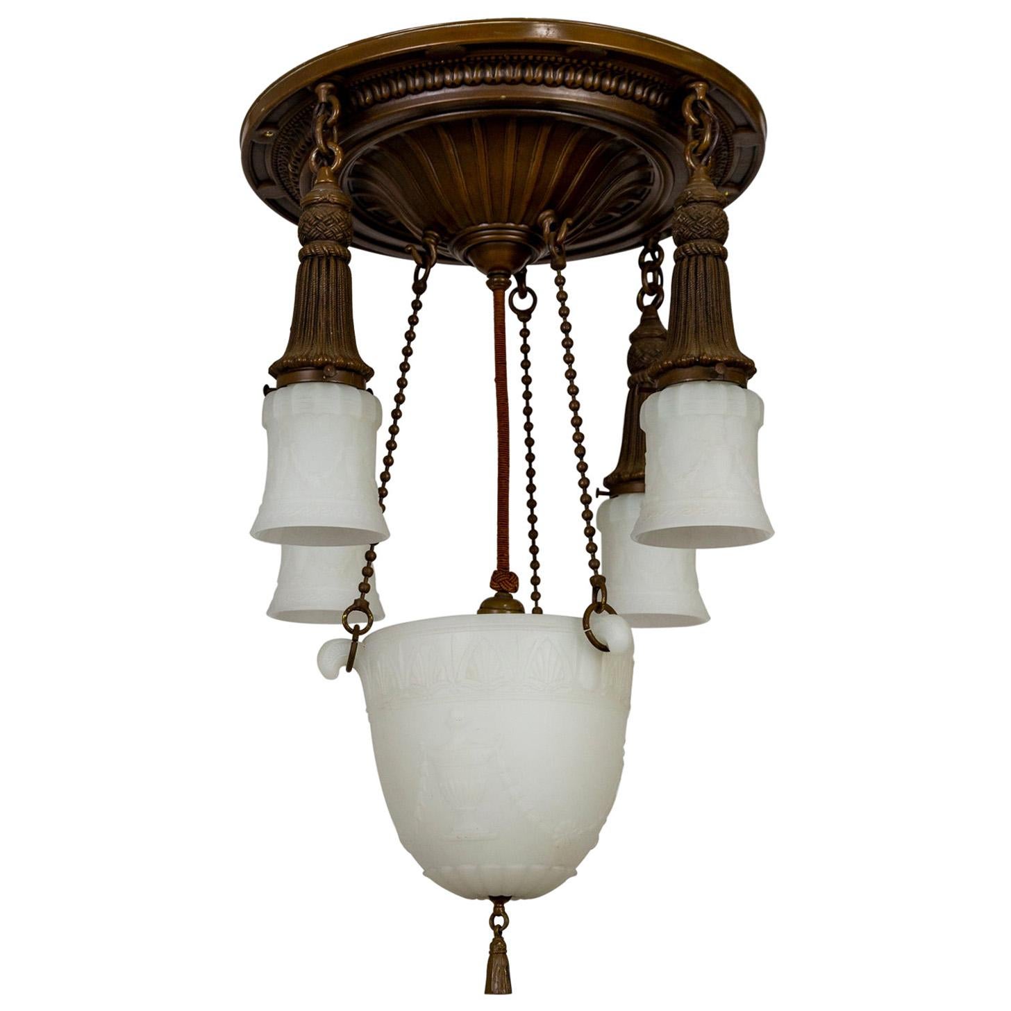An early 20th century, shower style chandelier with lacquered brass and milk glass. Details include beaded chains, shade holders in the form of tasseled ropes, and heavy, molded glass; with embossed urns, floral garlands, and Egyptian motifs; center