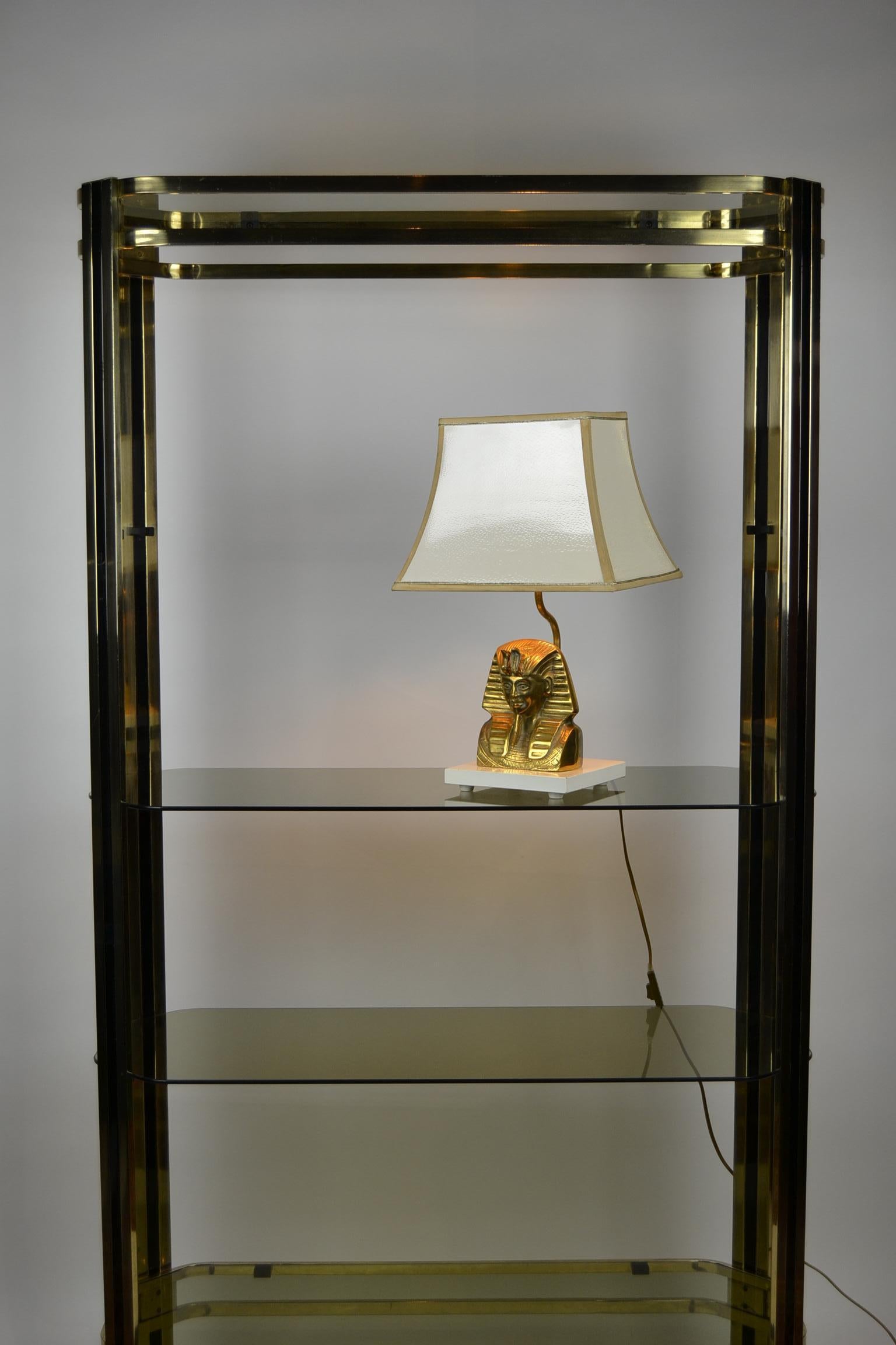 Pharaoh Head Table Lamp. 
This vintage table lamp or desk lamp dates from the 1970s.
It's a brass Pharaoh head sculpture on a white lacquered wooden base.
It has a white shiny lampshade which has the visual of an ostrich leather pattern.
It's gold