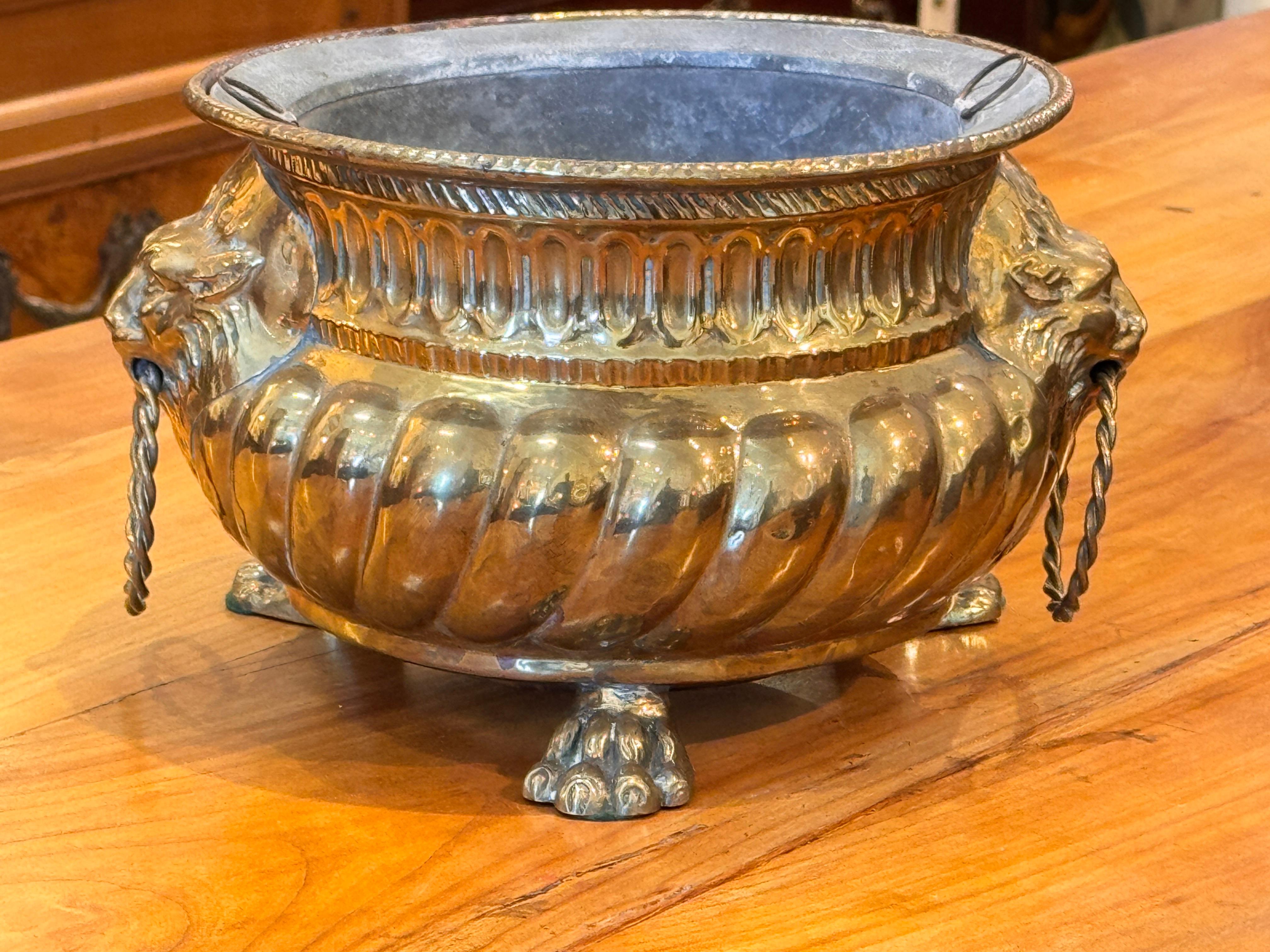 Brass planter with ornate repoussé foliate and scrollwork designs, set on three paw feet. Lion mask handles with rings flank each side, adding decorative appeal. Warm patina highlights the detailed craftsmanship.