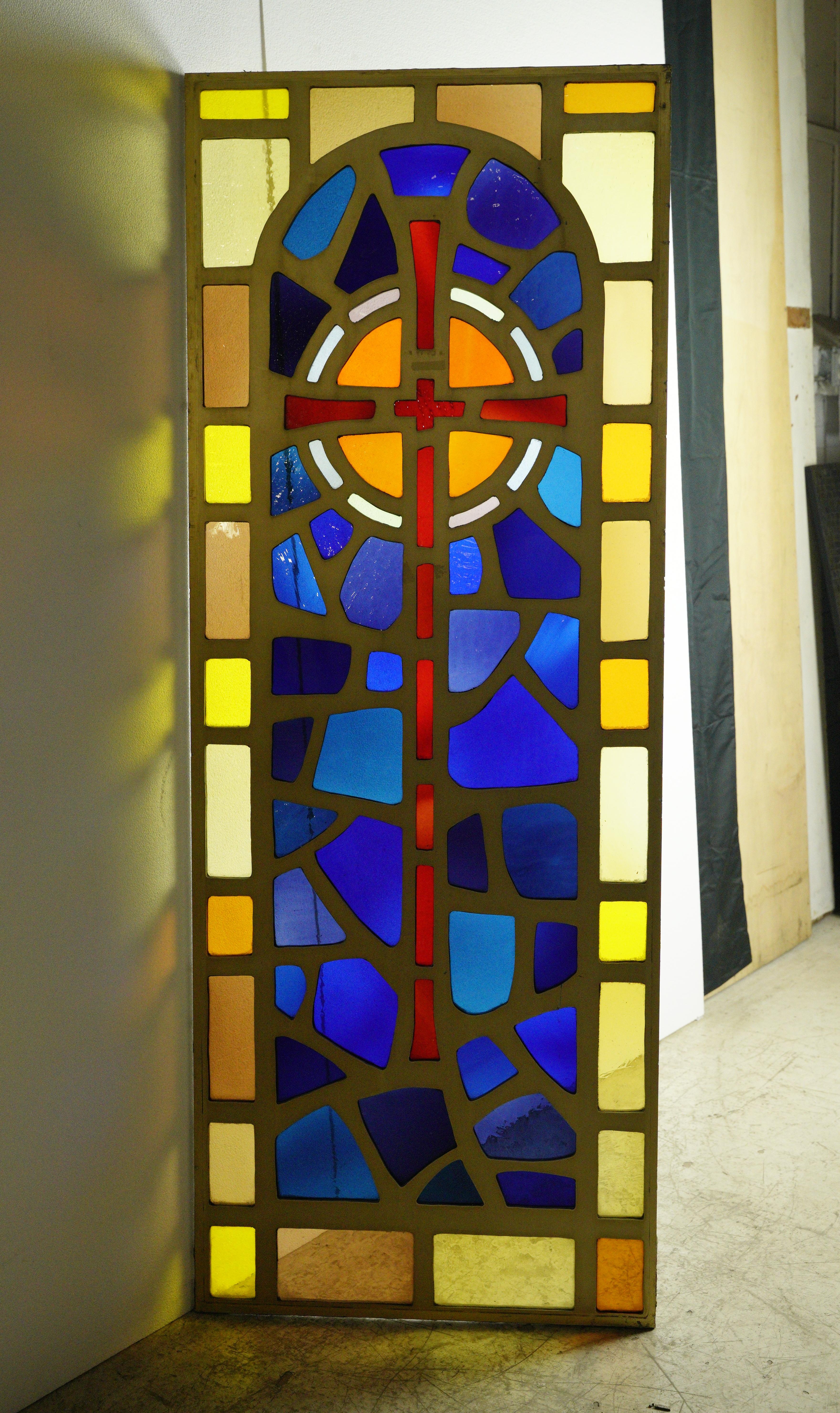 Brass coated aluminum church window with vibrant hues of blue, orange, and yellow stained glass with a centered red cross. Good condition with appropriate wear from age, with slight damage around the edges from its removal. One available. Please
