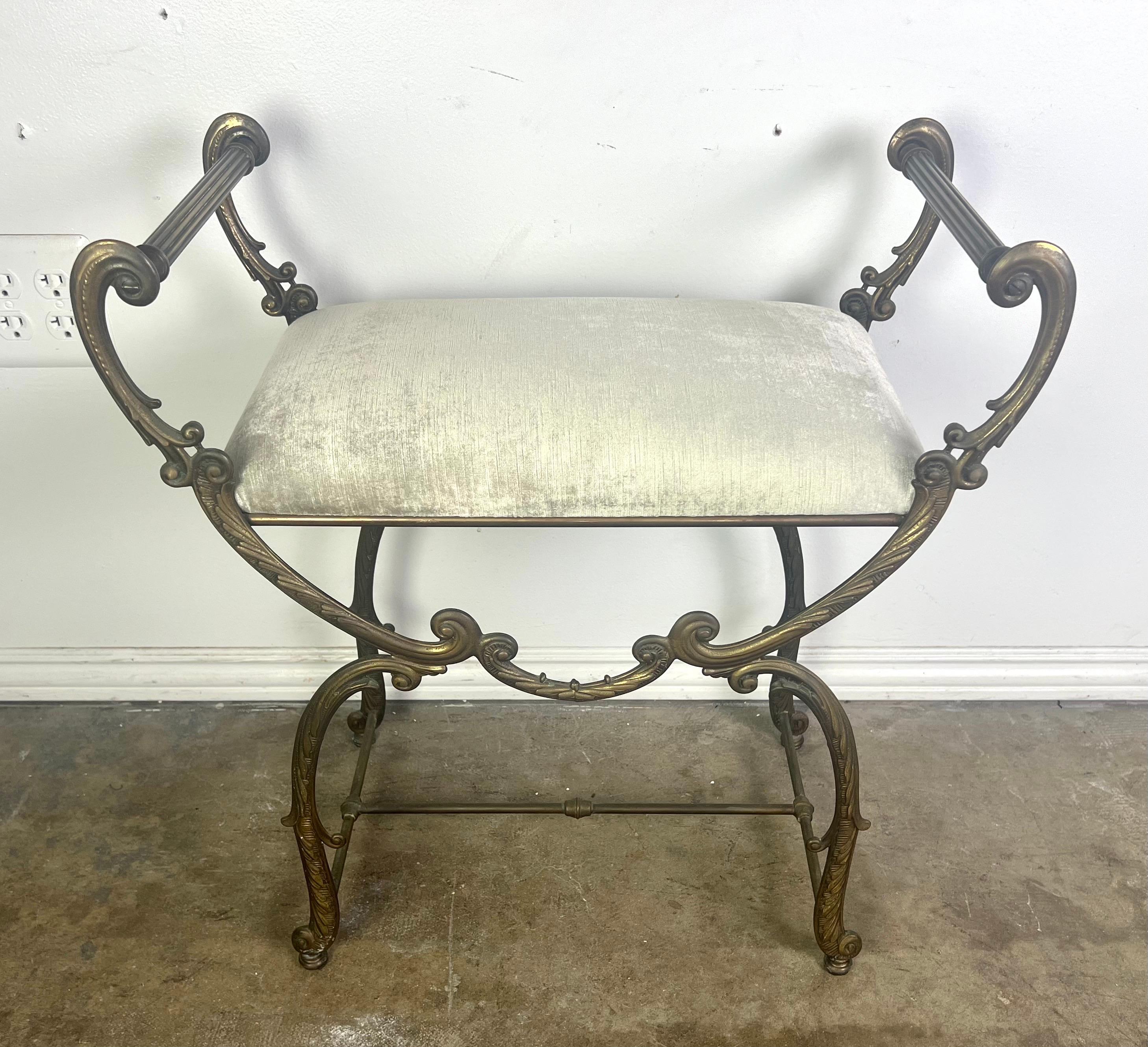 This French rococo style brass vanity bench features an ornate elegant design, showcasing a metal frame with intricate scrollwork and a luxurious antique finish.  The bench is upholstered in a silvery cream velvet that adds to it's opulent