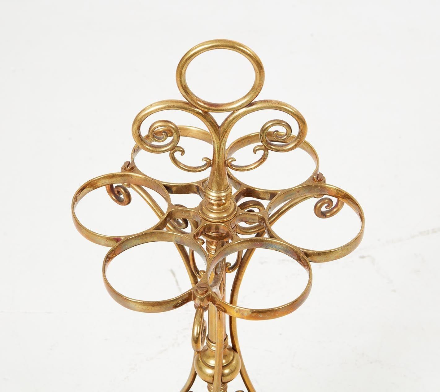 A scrollwork brass umbrella and/or walking-stick stand with ringed holders around a central shaft and three scrolled brackets surmounted by a conforming ring and top handle and ending in a heavy brass base with three sectional iron drip pan inserts.