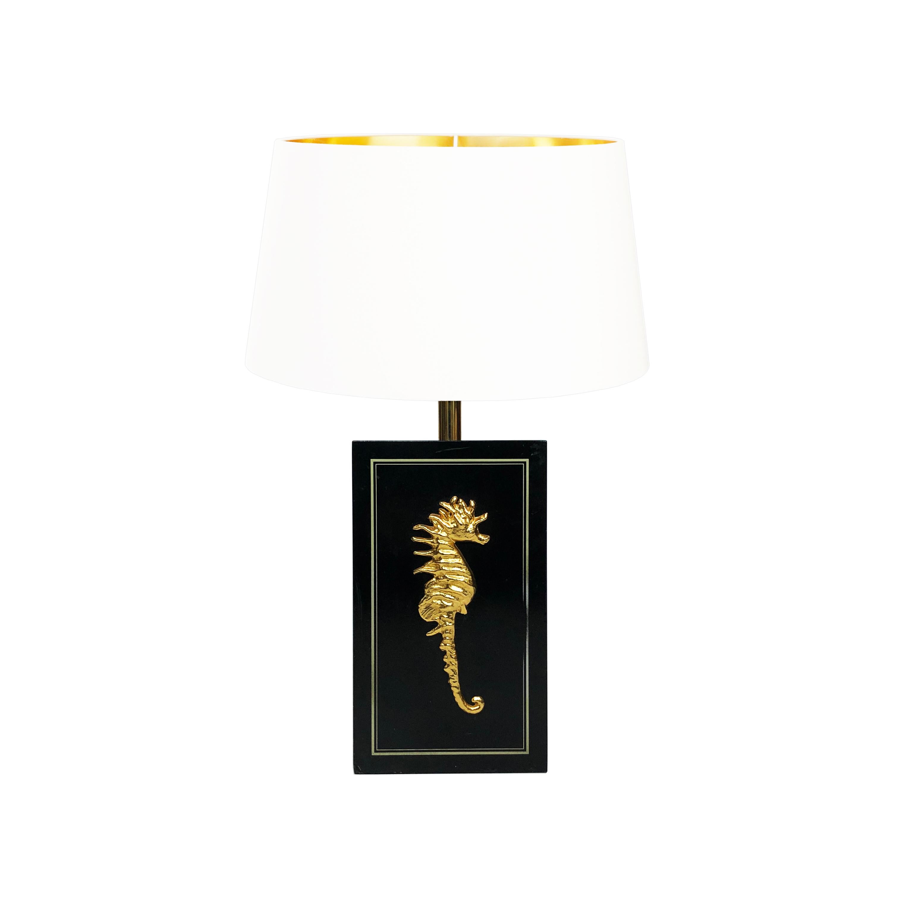 This Belgian table lamp in black and gold depicts the side profile of a seahorse, with an intricate brass plated hippocampus. This is mounted to a rectangular lacquered wood base, from which protrudes the bulb holder. There is a thin gold trim