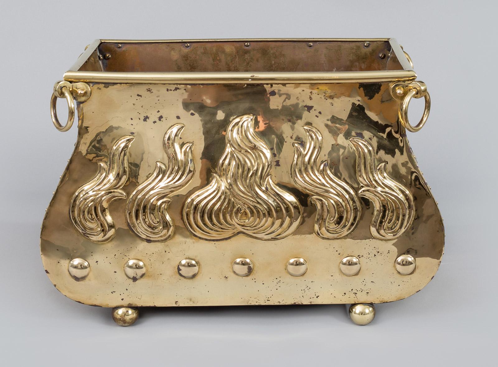 Brass coal scuttle with serpentine shaped sides, embossed flames on the front and back, attached to each corner is a brass ring, mounted on ball feet. Great for fire wood or kindling.