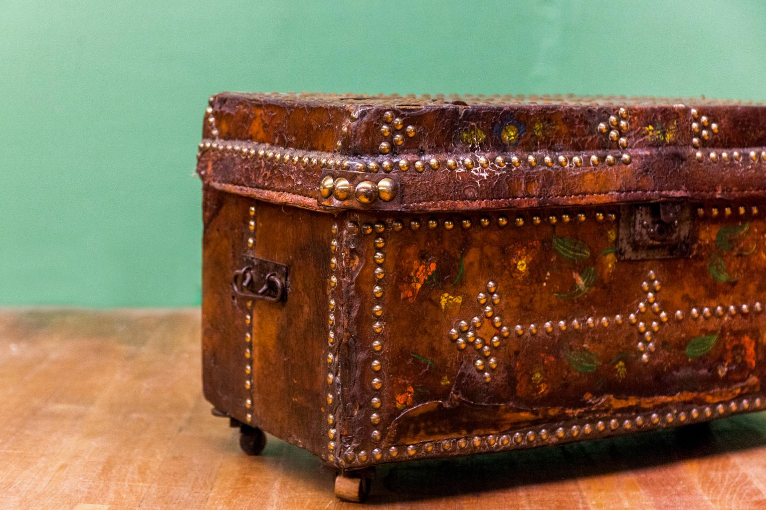 Brass studded painted leather box, has the original steel carrying handles, lock, and brass and wooden castors. There is a three inch long old shrinkage separation on the side.