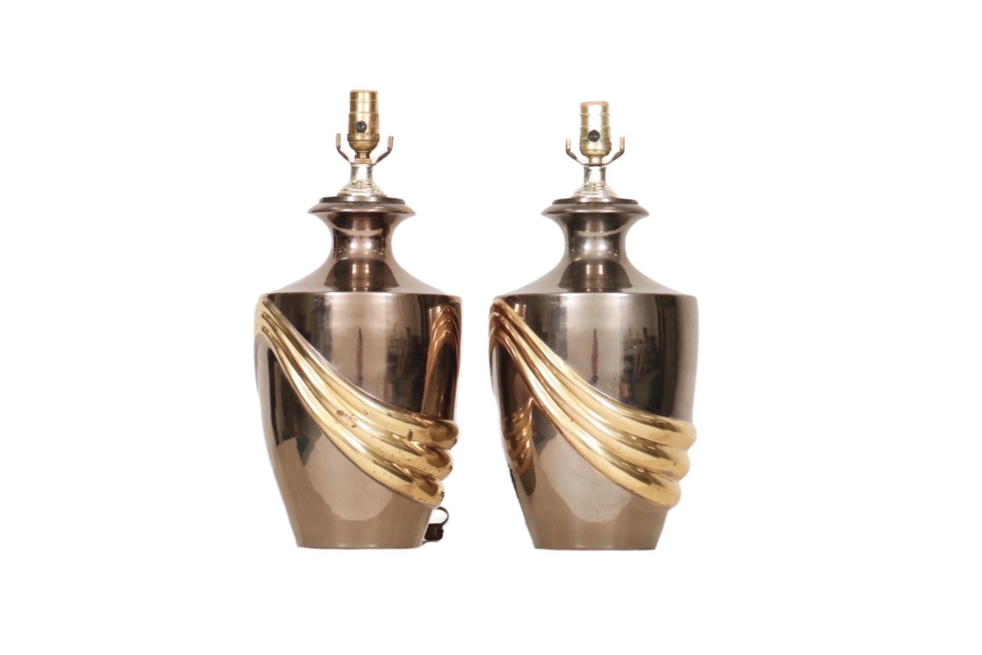 A large pair of Art Deco style urn shaped lamps made of brass, polished to a mirrored finish. Brass sockets are supported on beveled necks that curve down to angular shoulders. Embossed with an elegant swag motif in gold that drapes around the body