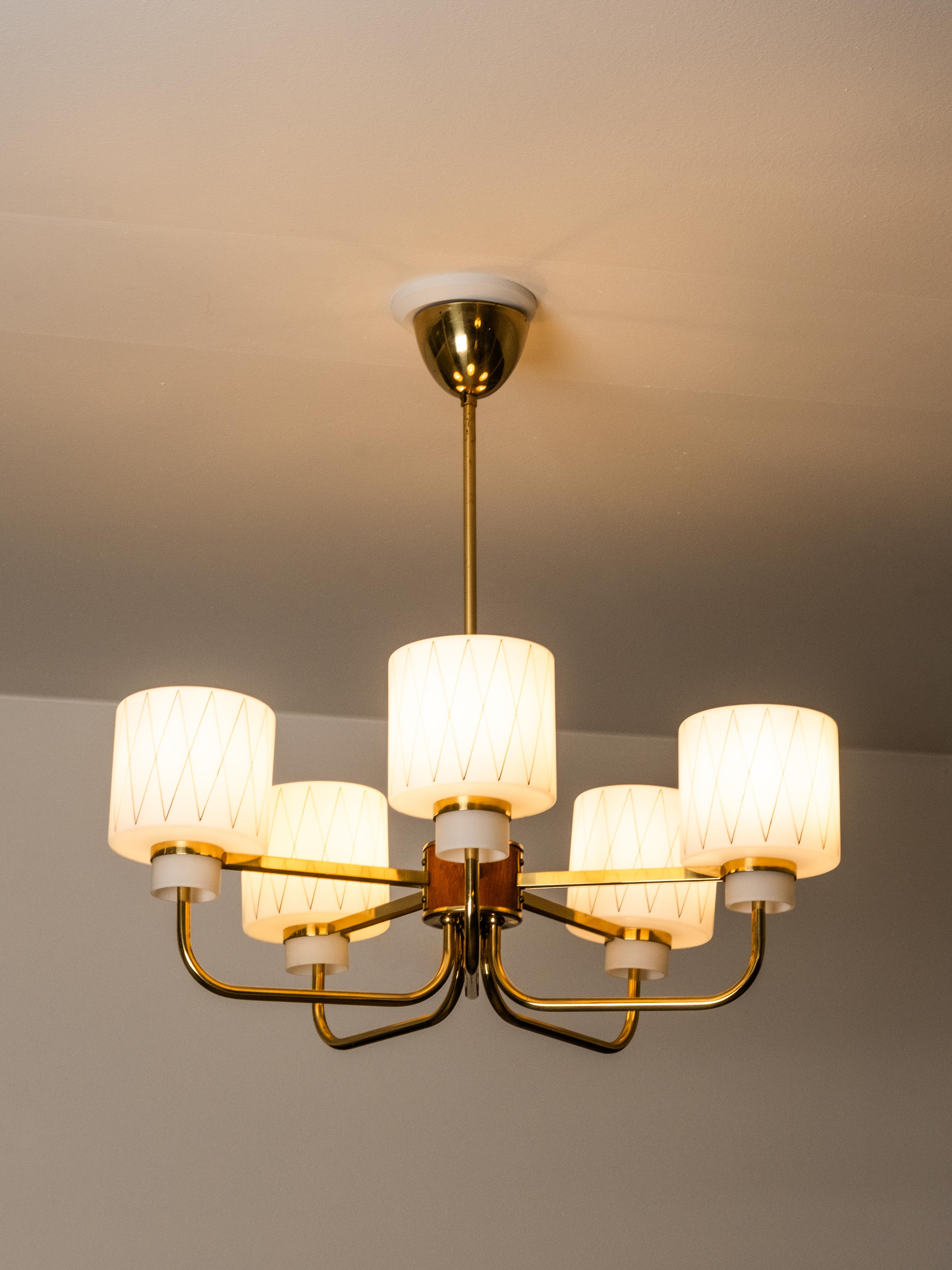 Un raffinato esempio di design finlandese di metà secolo. Questo lampadario a cinque bracci combina tubi in ottone con dettagli in teak e paralumi in vetro opalino, ciascuno decorato con un delicato motivo a diamante. La geometria equilibrata e la