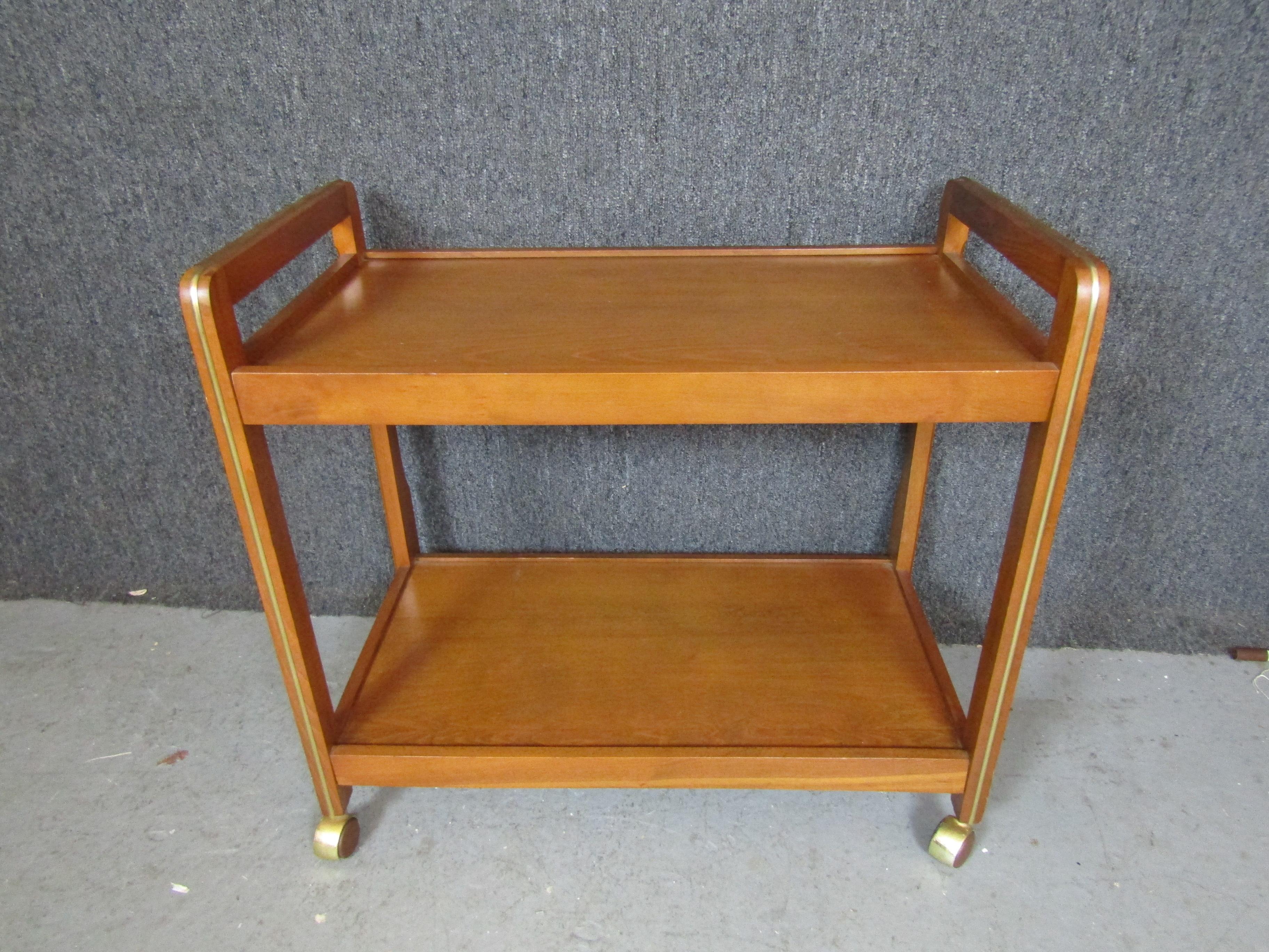 Mid-century modern rolling tea cart with brass trim around warm teak. Two shelves and wheels makes a great party bar or serving cart.
Please confirm location NY or NJ