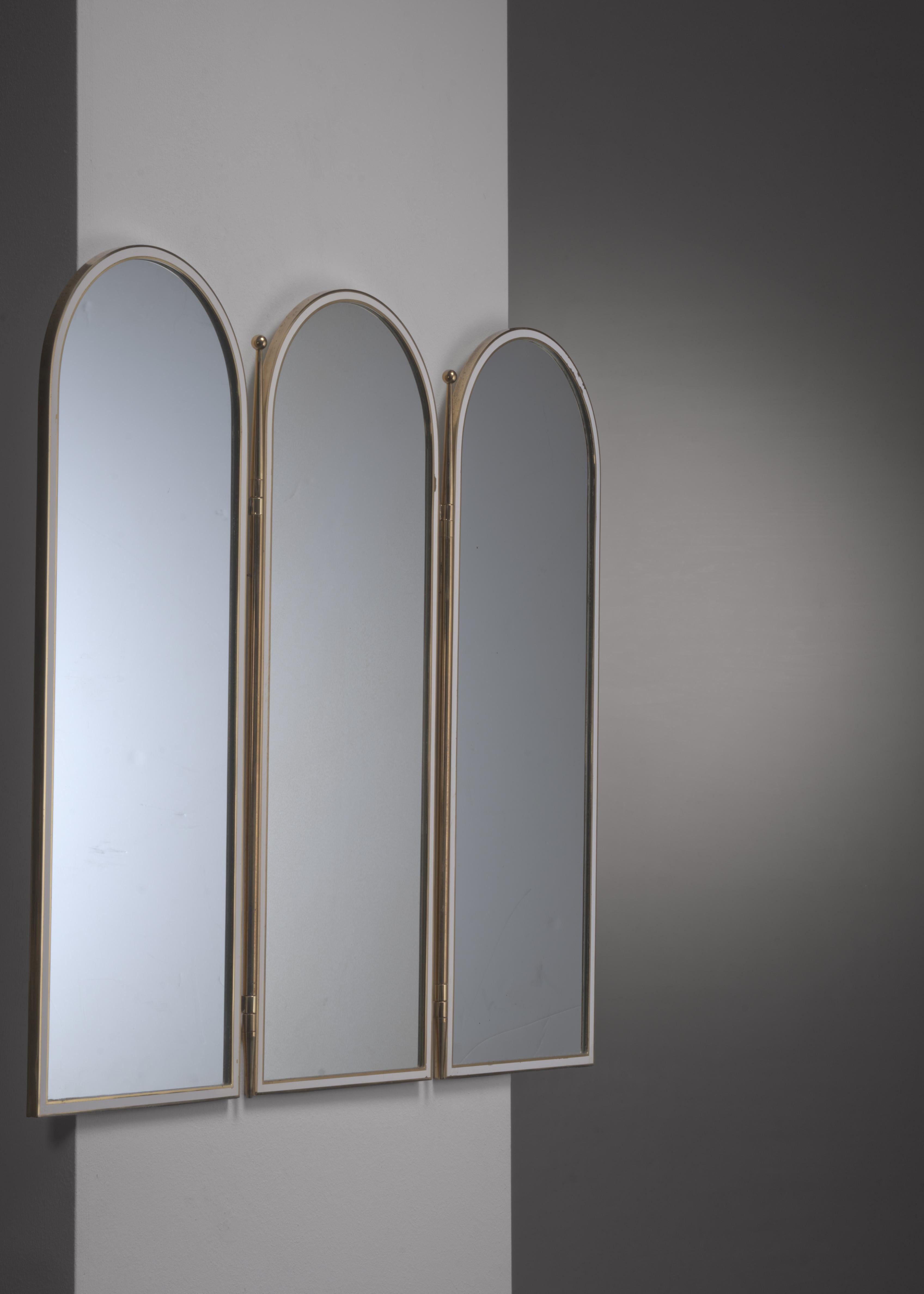 A triptych wall mirror in a brass frame with a white inner rim. The three separate mirror are connected with decorative brass hinges.