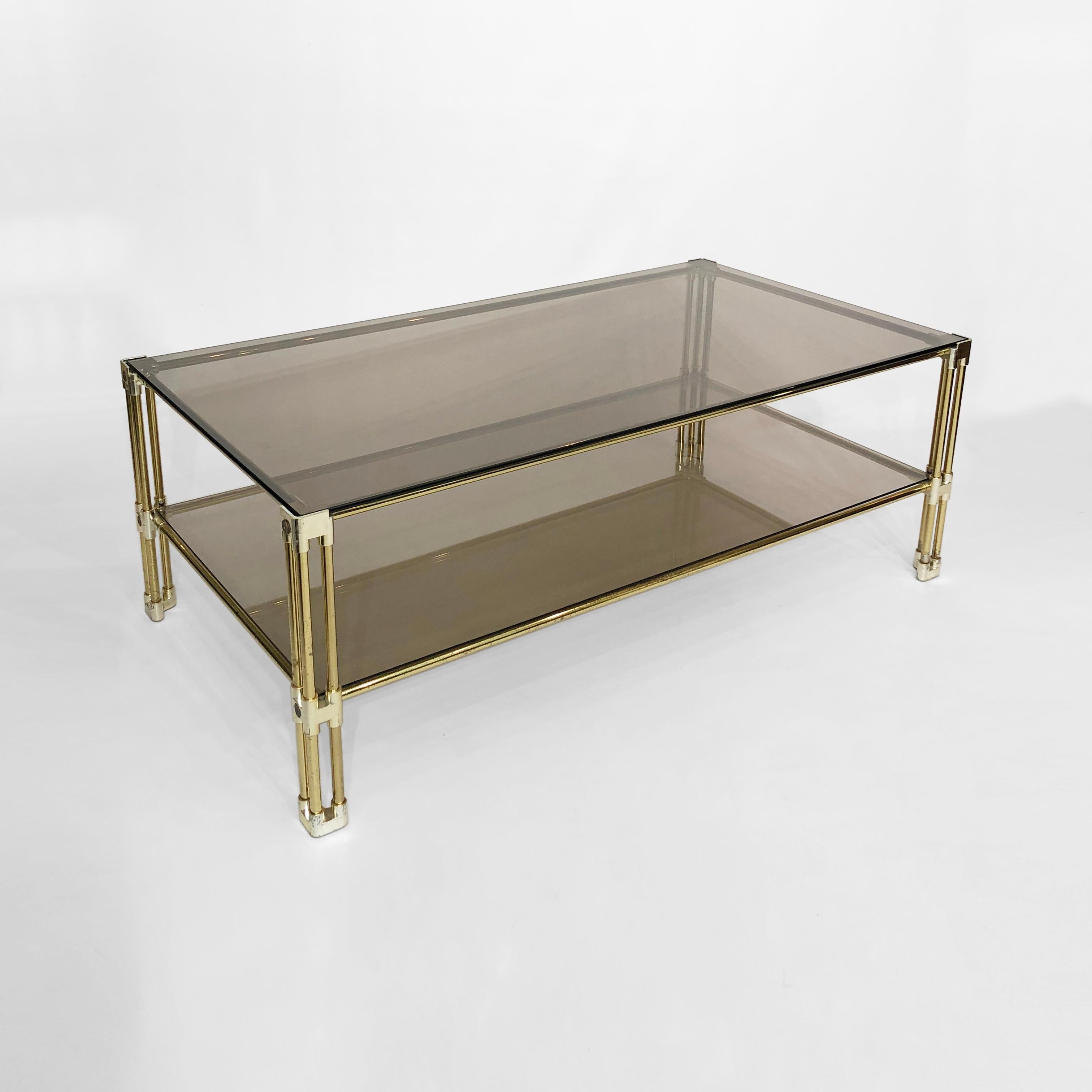 A two-tiered brass and smoked glass coffee table, believed to have originated in 1970s Italy. This prime example of Hollywood Regency design features two sheets of tempered smoked glass, housed in a brass frame. The corners and joints of the frame