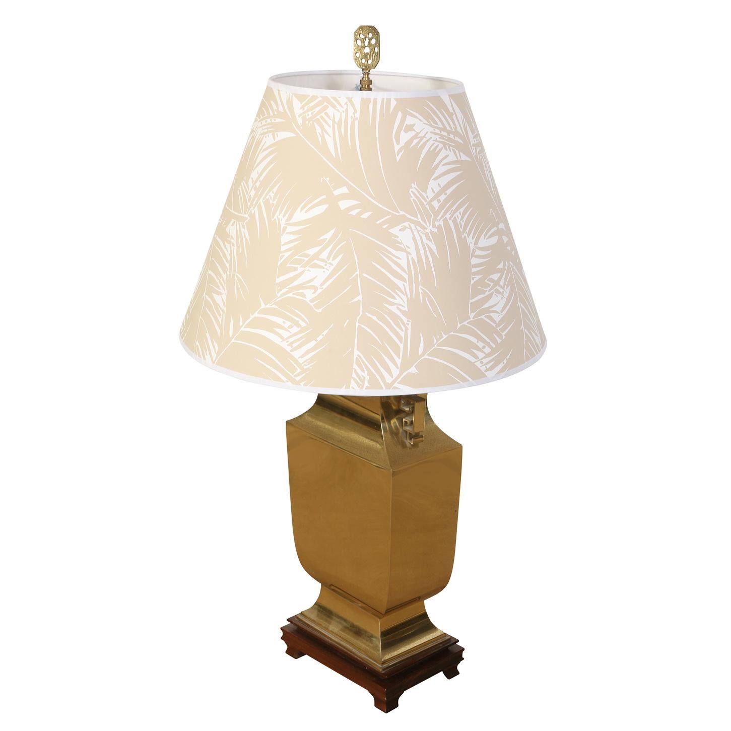 A brass urn lamp with fret chinoiserie motif handles on a wood base. The lamp's body is crafted from lustrous brass, giving it a rich, golden hue that catches the light beautifully. However, the brass has developed some pitting over time, adding