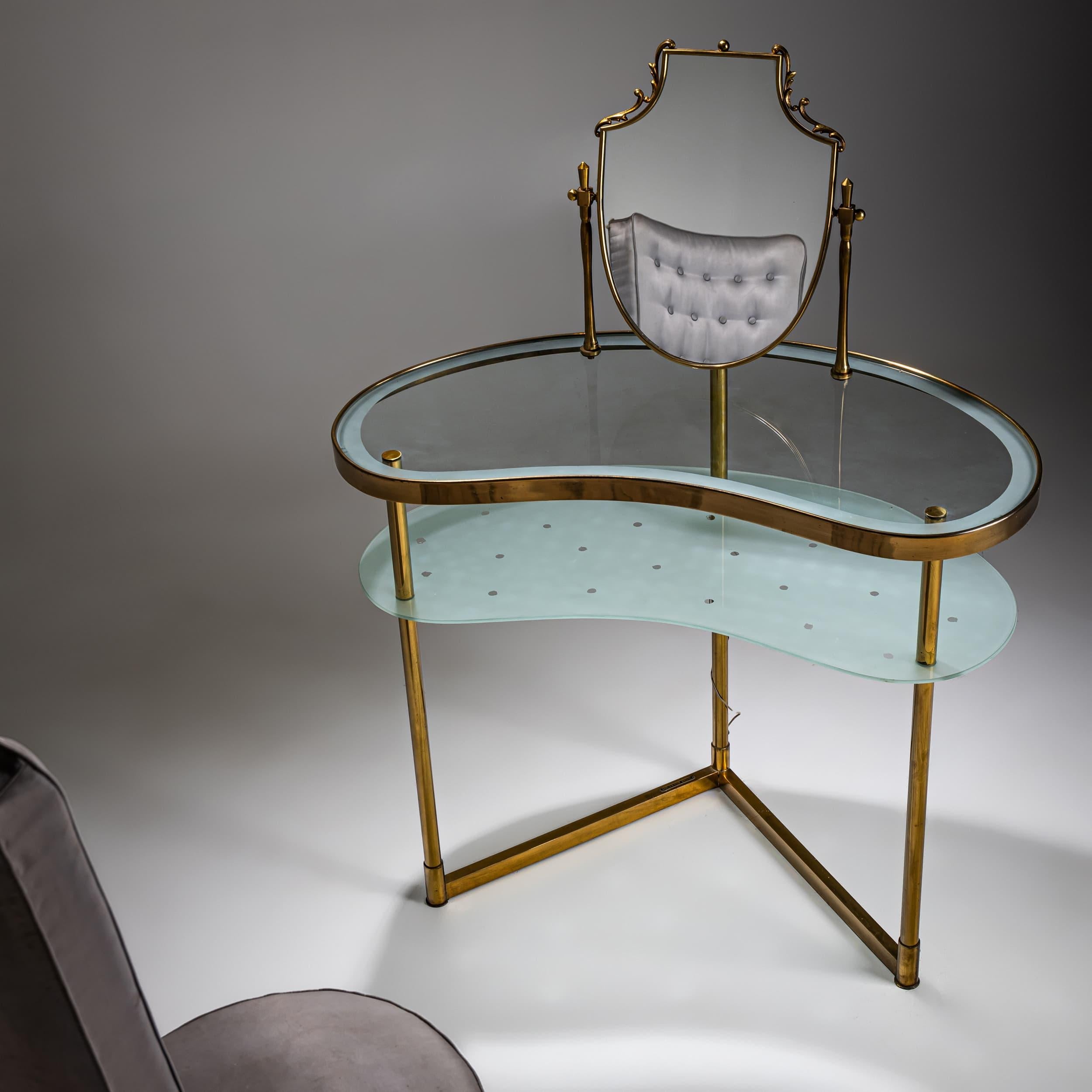 Élégante coiffeuse en laiton de forme réniforme, dotée de deux surfaces en verre, partiellement dépoli et orné d'un motif en forme d'étoile. Le miroir en forme de bouclier est monté sur un cadre pivotant, tandis que la table est éclairée par le bas,