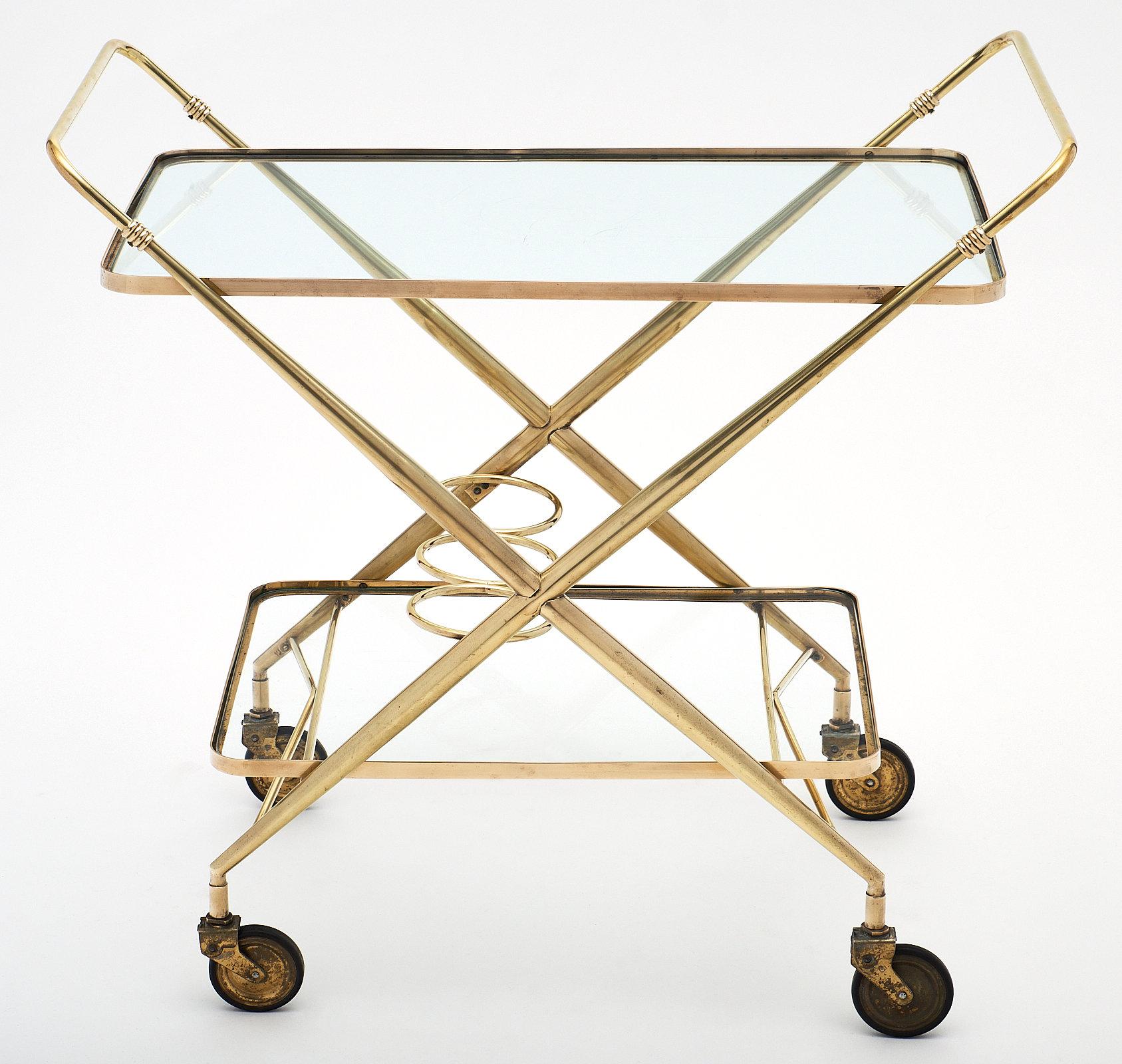 French vintage brass bar cart on casters with two glass shelves and three bottle holders on the bottom shelf. The x-shape of the profile and double-handle feature make this a sleek and functional design solution.