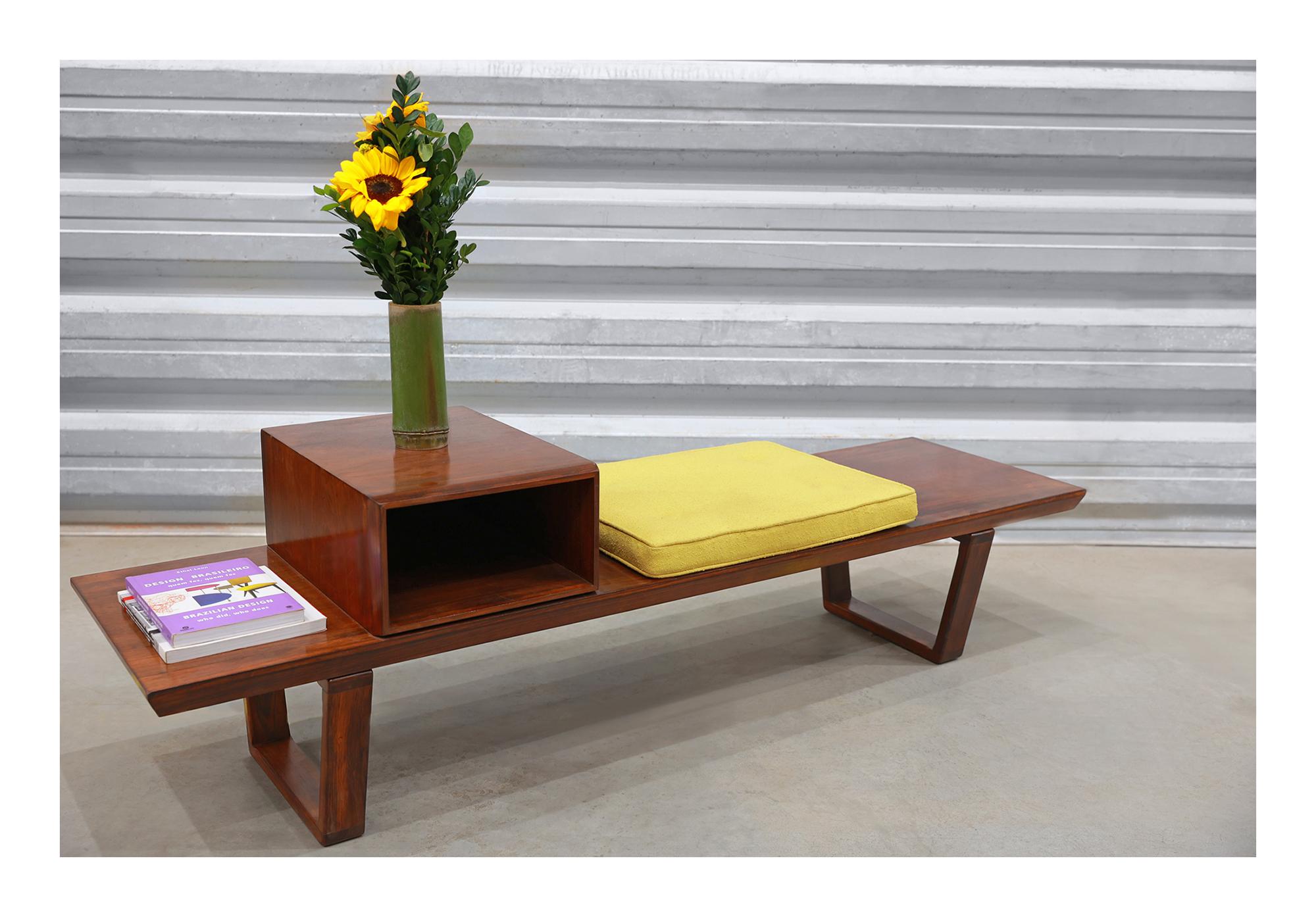 La banquette comporte deux pieds, un module pour le rangement et une assise avec un tissu jaune retapissé. La pièce est fabriquée en bois dur brésilien et présente une couleur et un motif magnifiques. Le bois a également été remis à neuf et est en