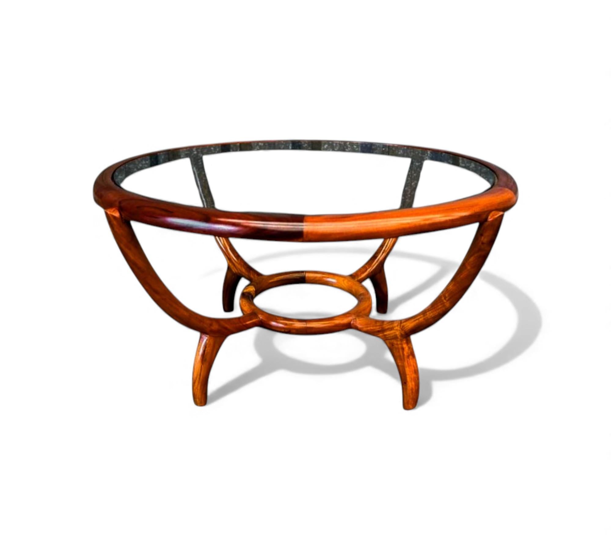 L'étonnante table basse ronde conçue par Giuseppe Scapinelli se caractérise par une base sculpturale en bois dur massif aux courbes fluides et aux formes entrecroisées. Le cadre circulaire supporte un plateau en verre, soulignant l'élégance