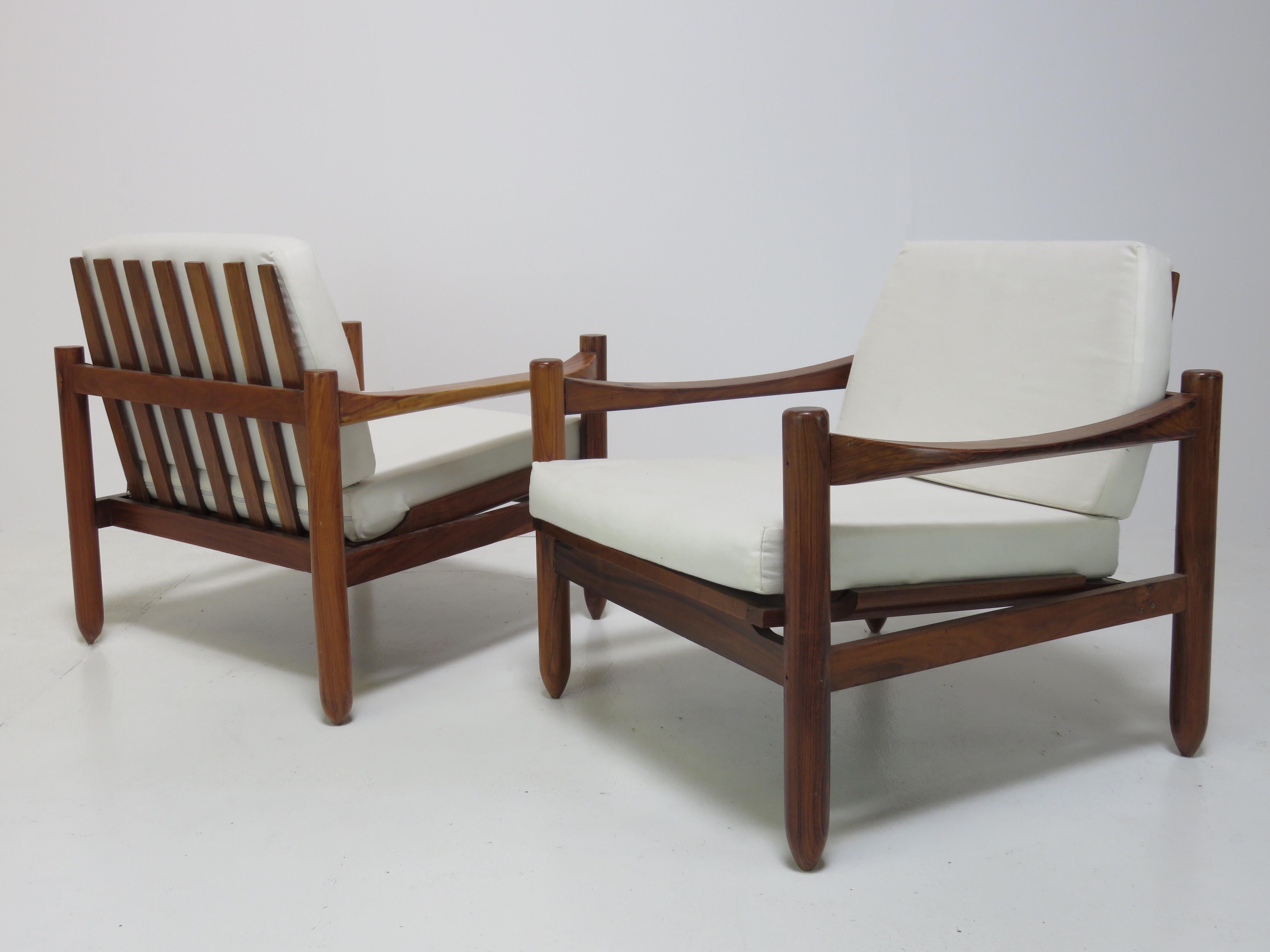 A pair of Brazilian hardwood lounge chairs. Attractive slatted back, cyclindrical legs and curved arms. Includes loose cushions covered in a white fabric.