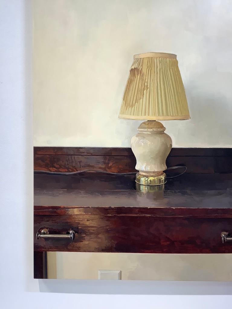 In this quiet still life painting, a single lamp with a yellowed lamp shade stands atop a dark brown mahogany wooden desk with gray metal handles on the drawer. The object and furniture is unexpectedly dramatic against a stark off-white wall; a